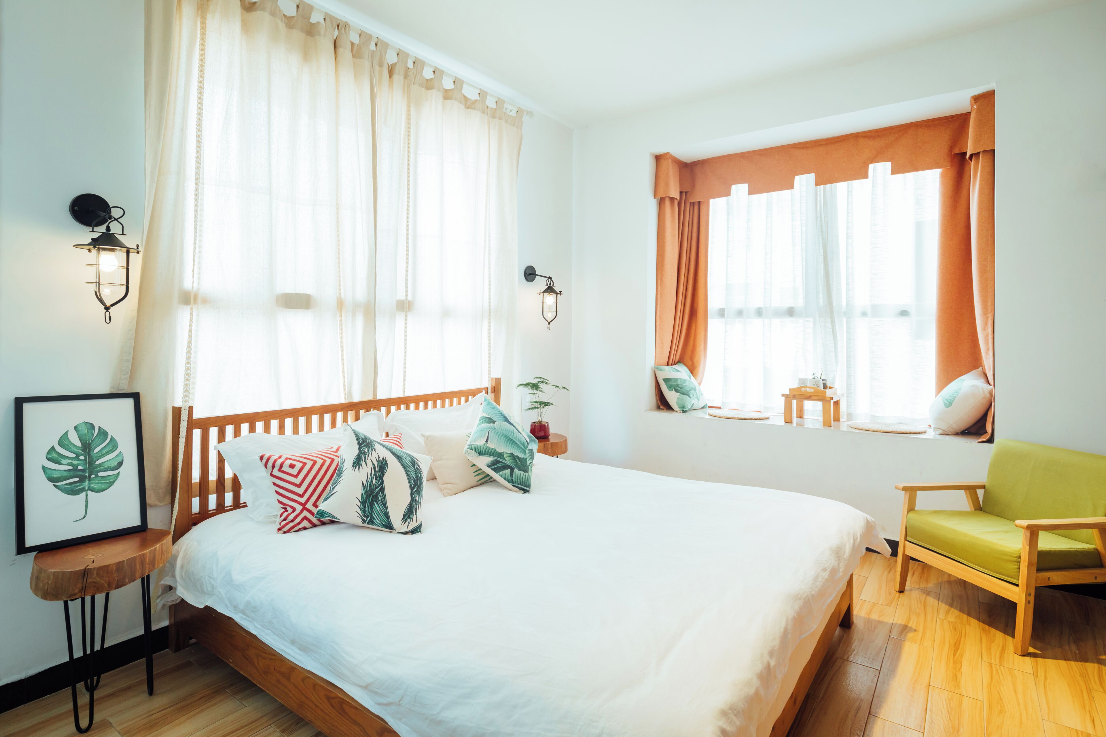A bedroom with a wooden bed, a green chair, a window with curtains, and wall-mounted lights.