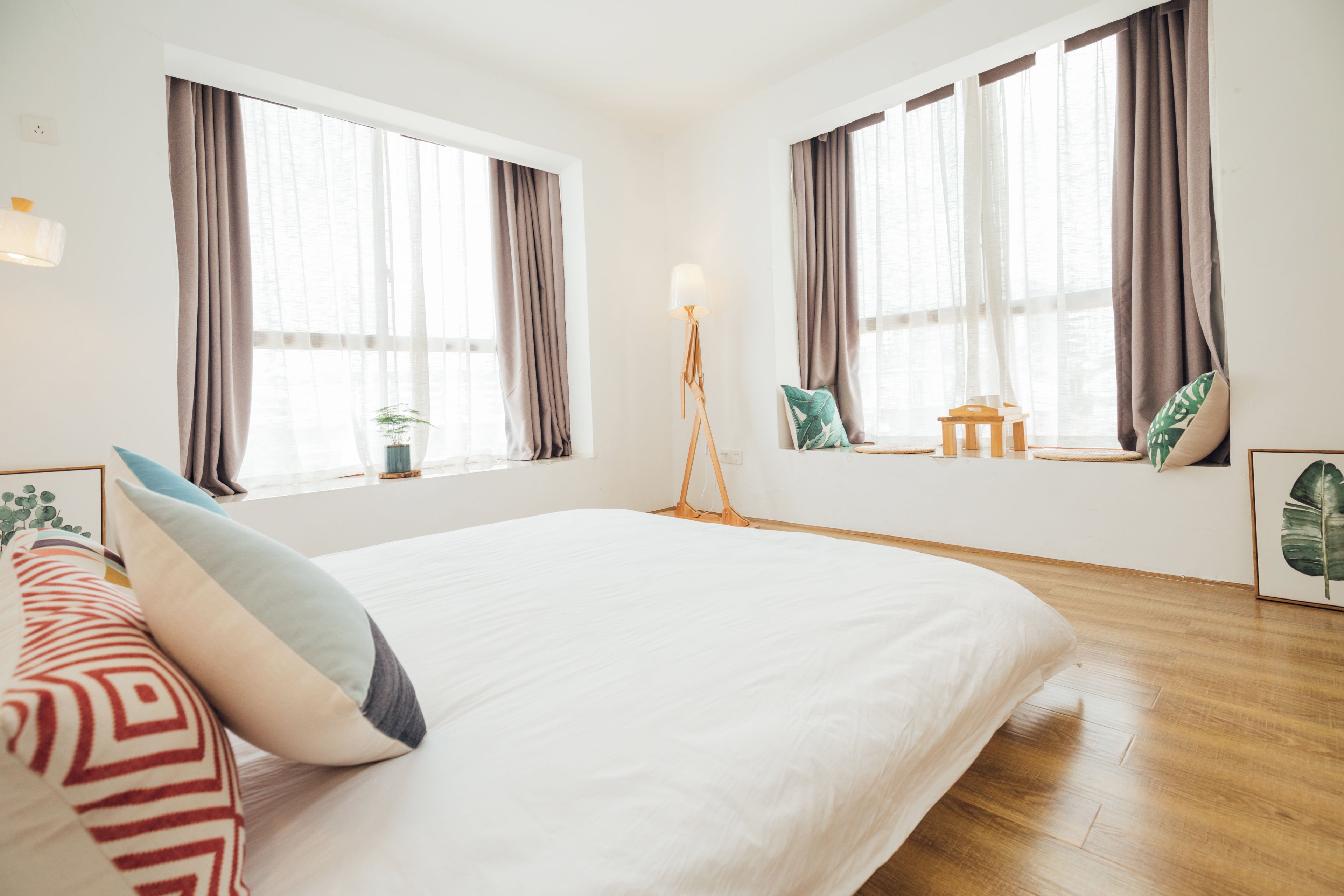 Habitación de hotel con una cama grande, dos ventanas con cortinas, una mesita de noche con lámpara y una pequeña planta.