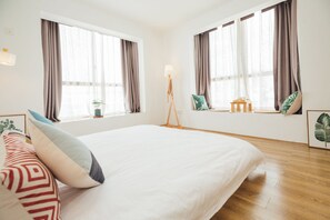 Hotellihuone, jossa on suuri sänky, kaksi ikkunaa, joissa on verhot, yöpöytä lampun kanssa ja pieni huonekasvi.