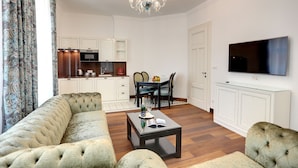 A living room with a sofa, a coffee table with a book and a bottle, a dining area with a table and chairs, a kitchen with cabinets and appliances, and a TV on the wall. - Ferdinandhof Apart-Hotel (Karlovy Vary)