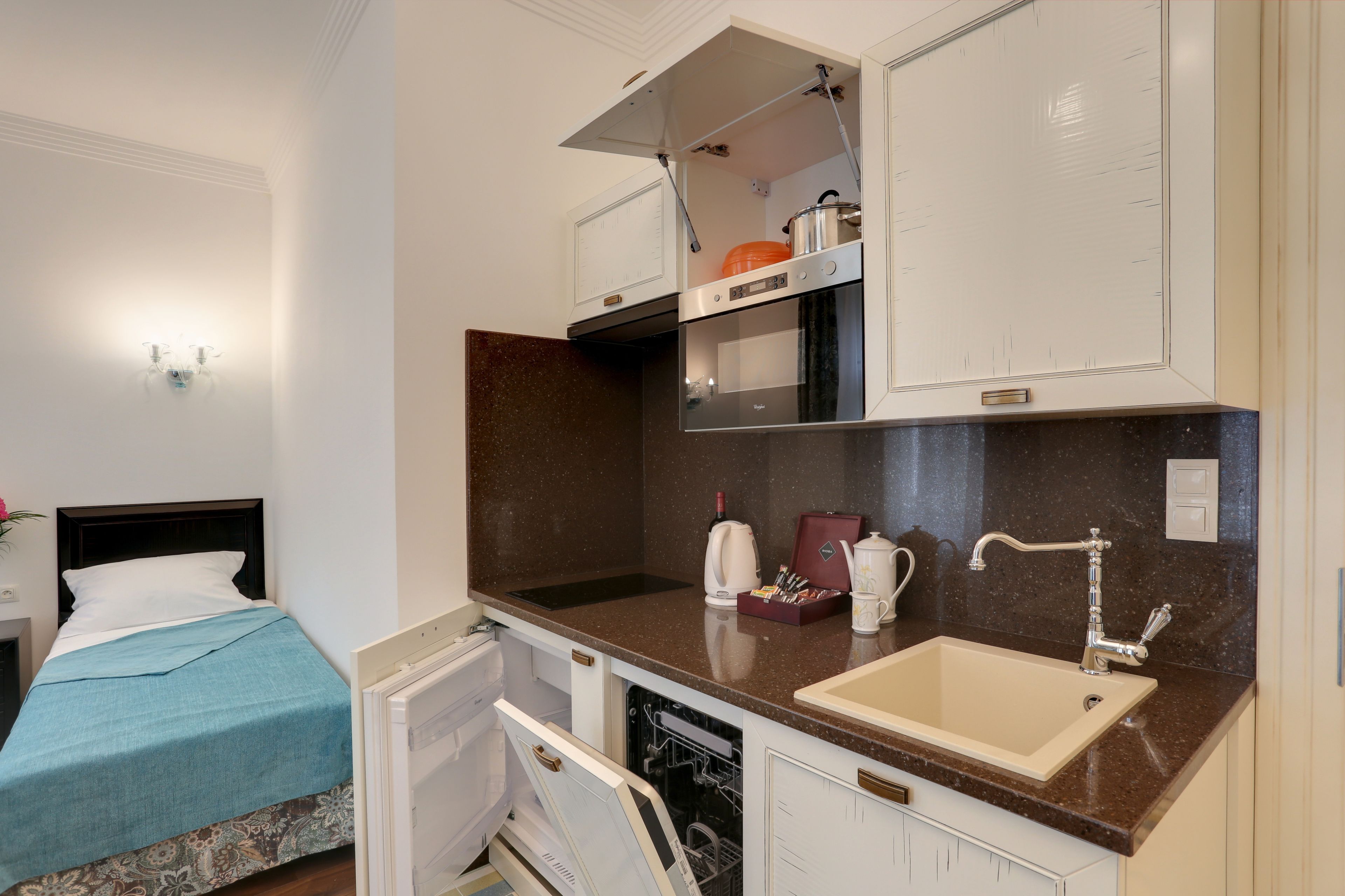 A compact kitchen with a built-in oven, a sink, and a microwave. There is a bed with a blue blanket and a white pillow in the adjacent room.