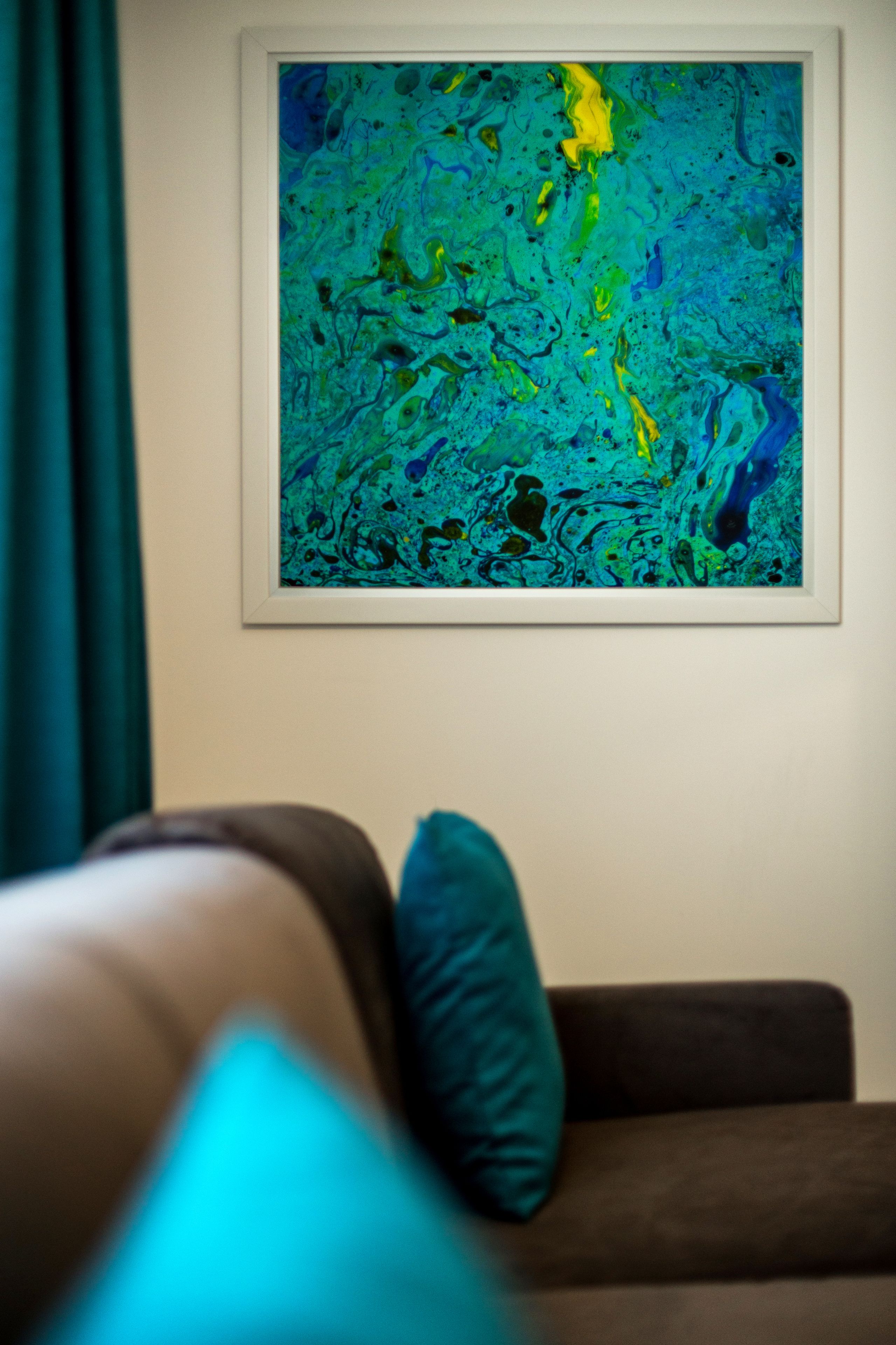 A framed abstract painting on a wall above a sofa with a teal cushion.