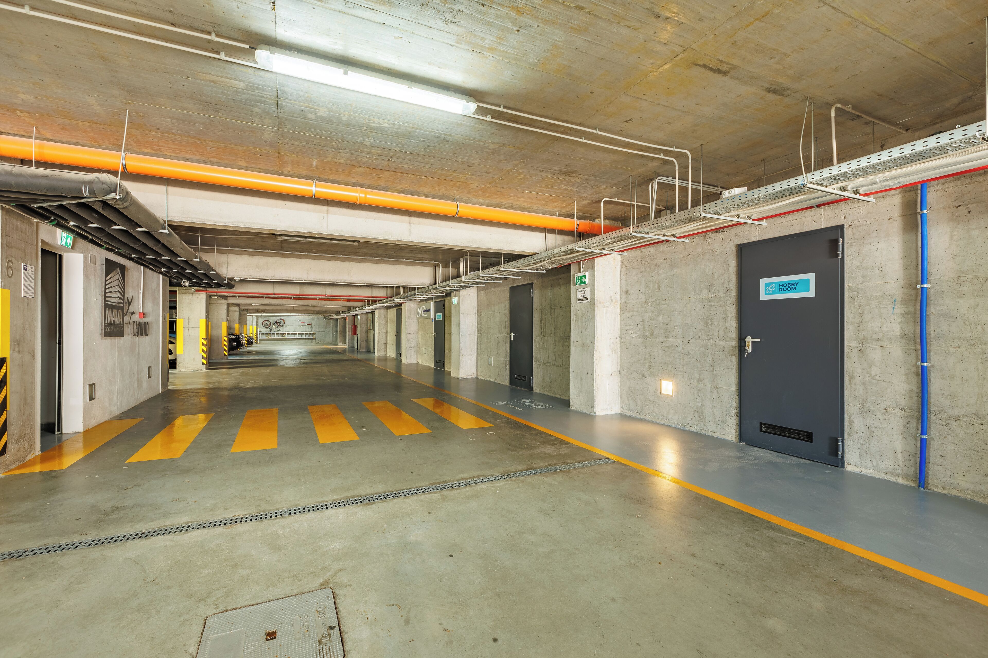 A clean, well-lit parking garage with concrete walls, yellow safety lines, and a blue door labeled "Hobby Room".