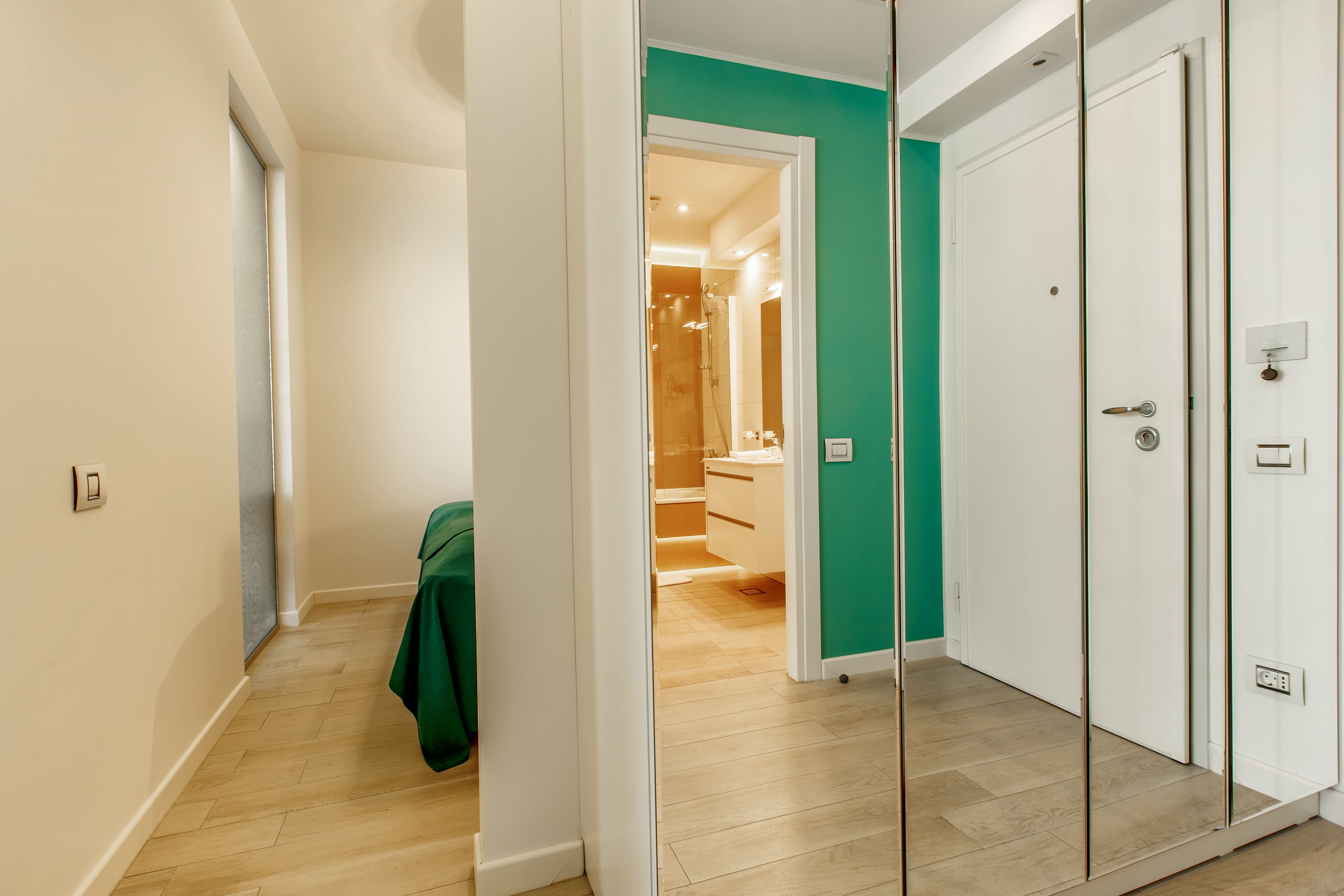 A hallway with a green accent wall, wooden flooring, and a door leading to a bathroom with a shower and a sink.