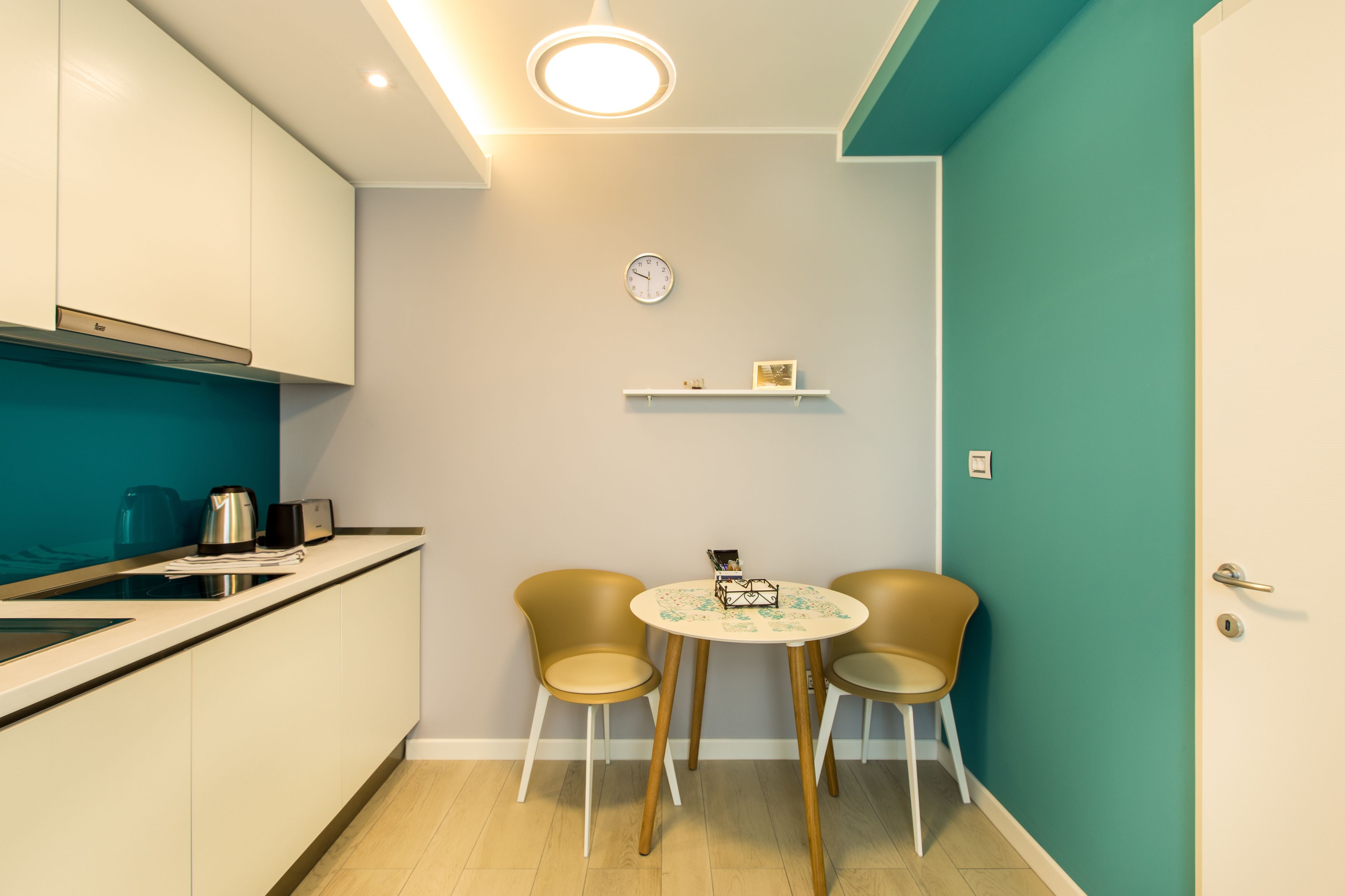 A compact kitchen with a dining area, including a table with chairs, a clock, and a shelf with decorative items.