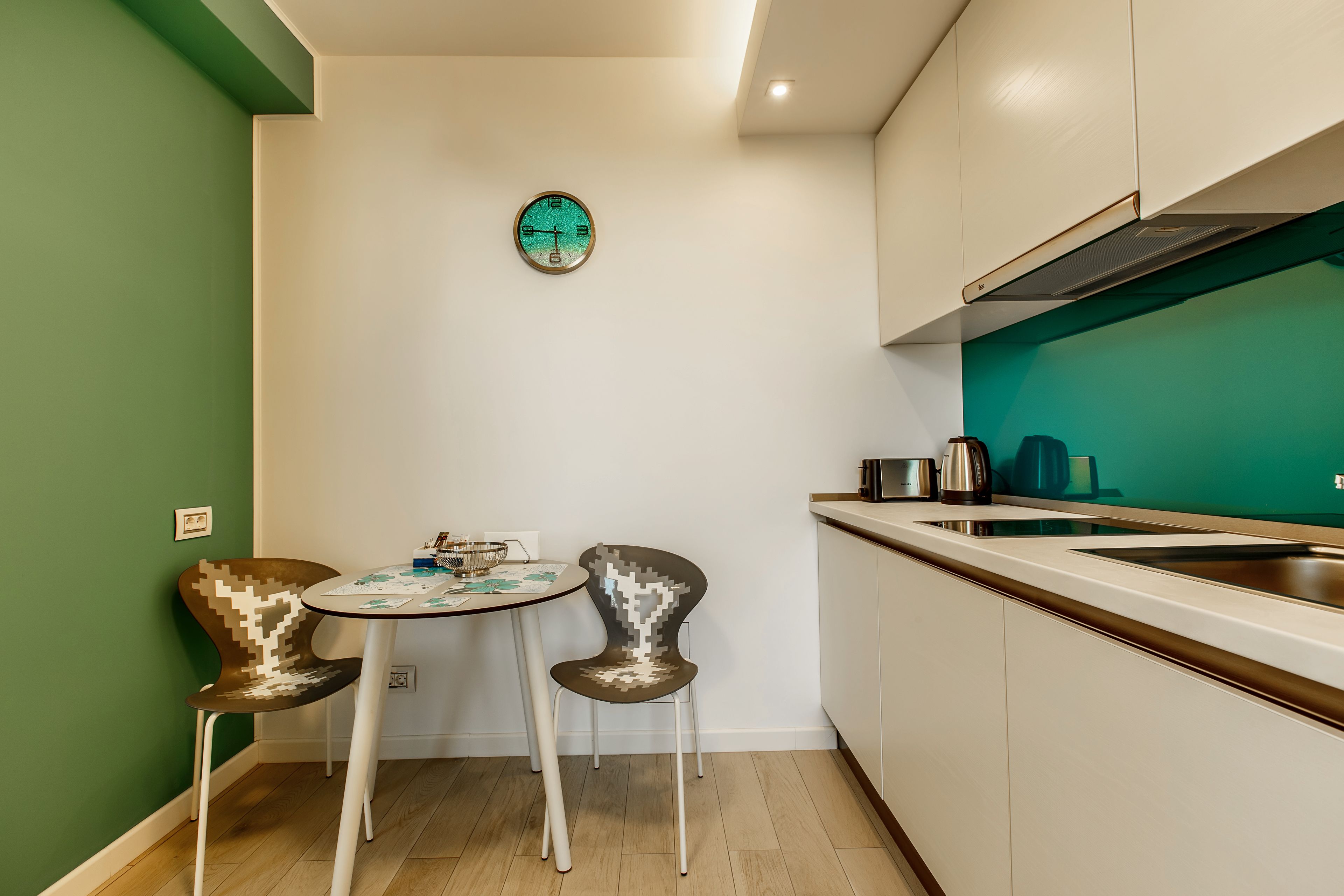 A compact kitchen with a dining area, a clock on the wall, and a green accent wall.
