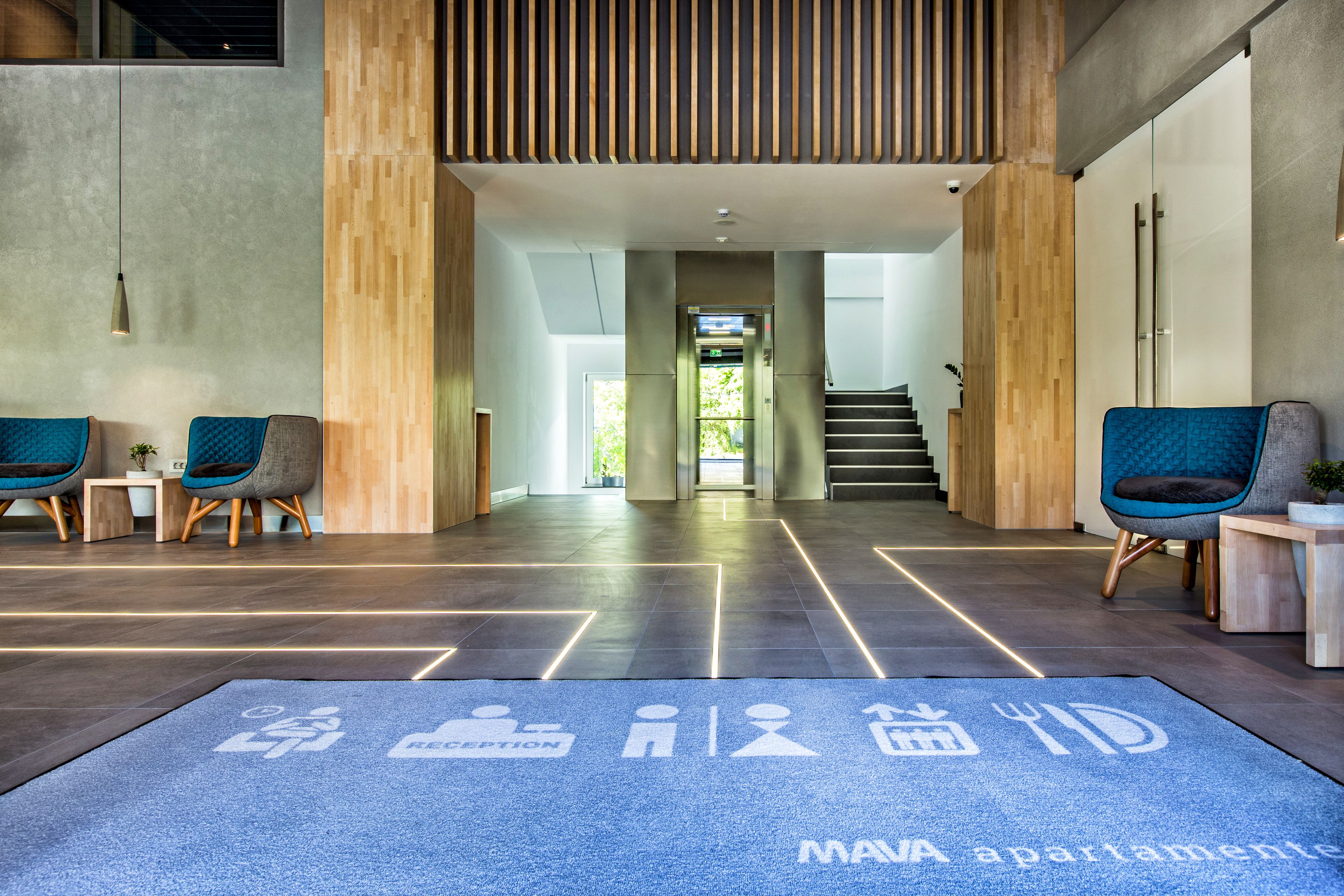 A modern lobby with a reception desk, two blue chairs, a blue carpet with icons, and a staircase leading to an upper level.
