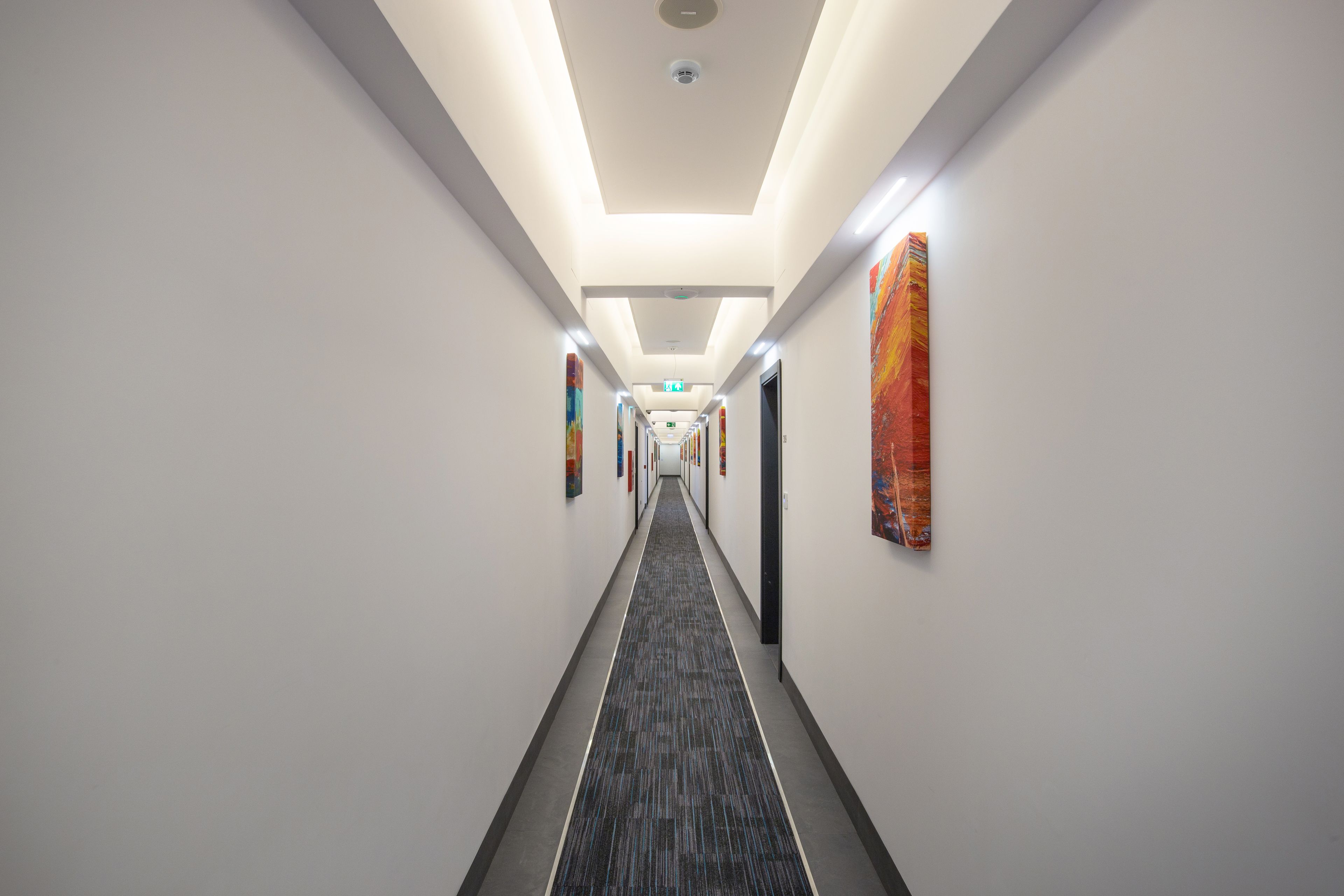 Un couloir d’hôtel moderne avec un sol recouvert de moquette, des murs blancs et des œuvres d’art accrochées aux murs.