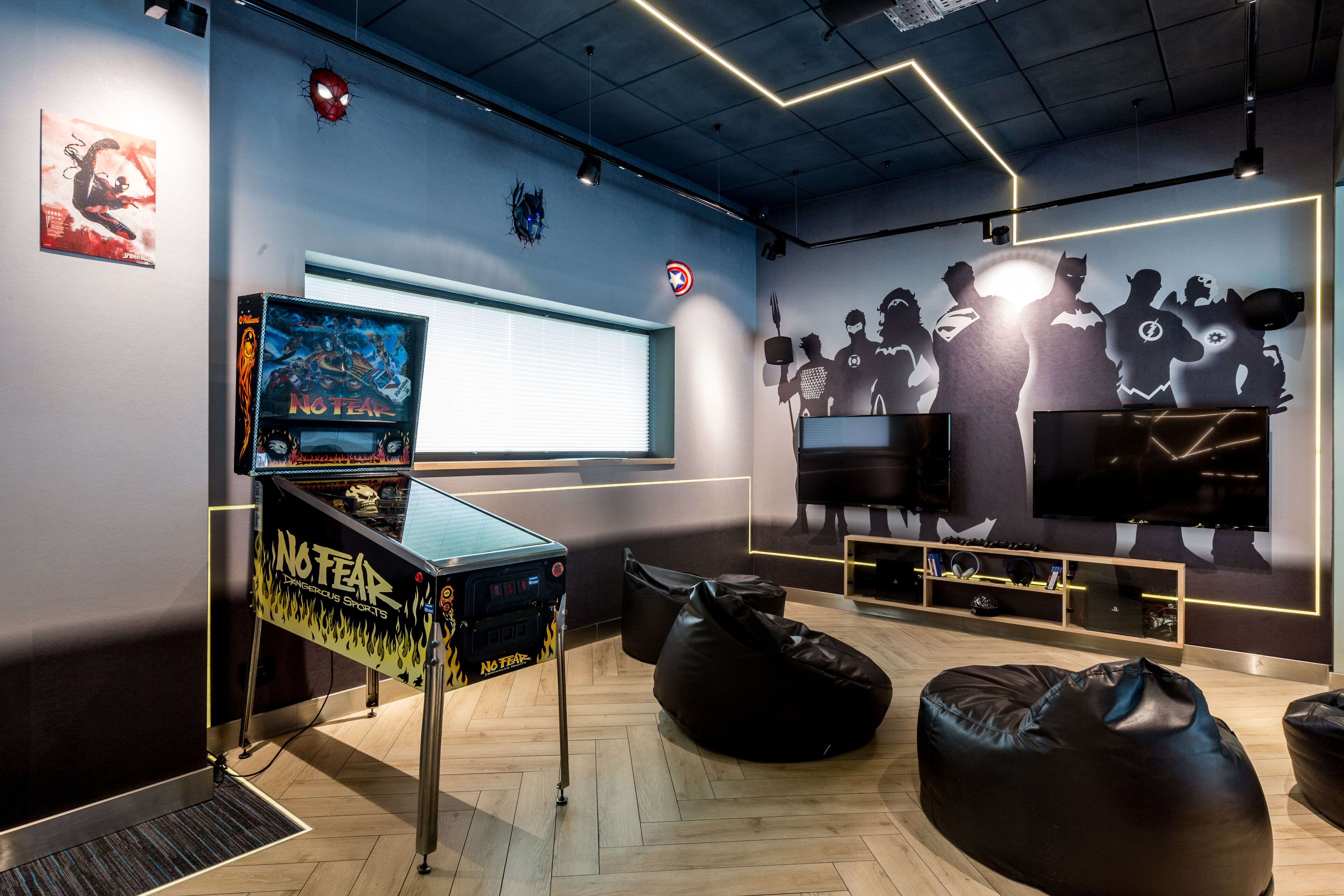 Una sala de juegos con una pantalla grande, sillones tipo bean bag y un mural de superhéroes.