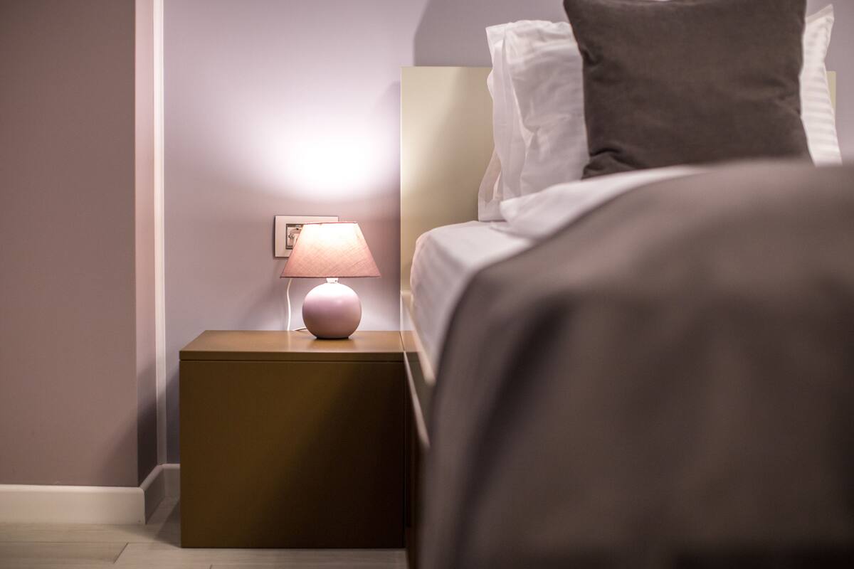 A neatly made bed with a white and gray pillow, a small bedside table with a pink lamp, and a wall-mounted picture frame.