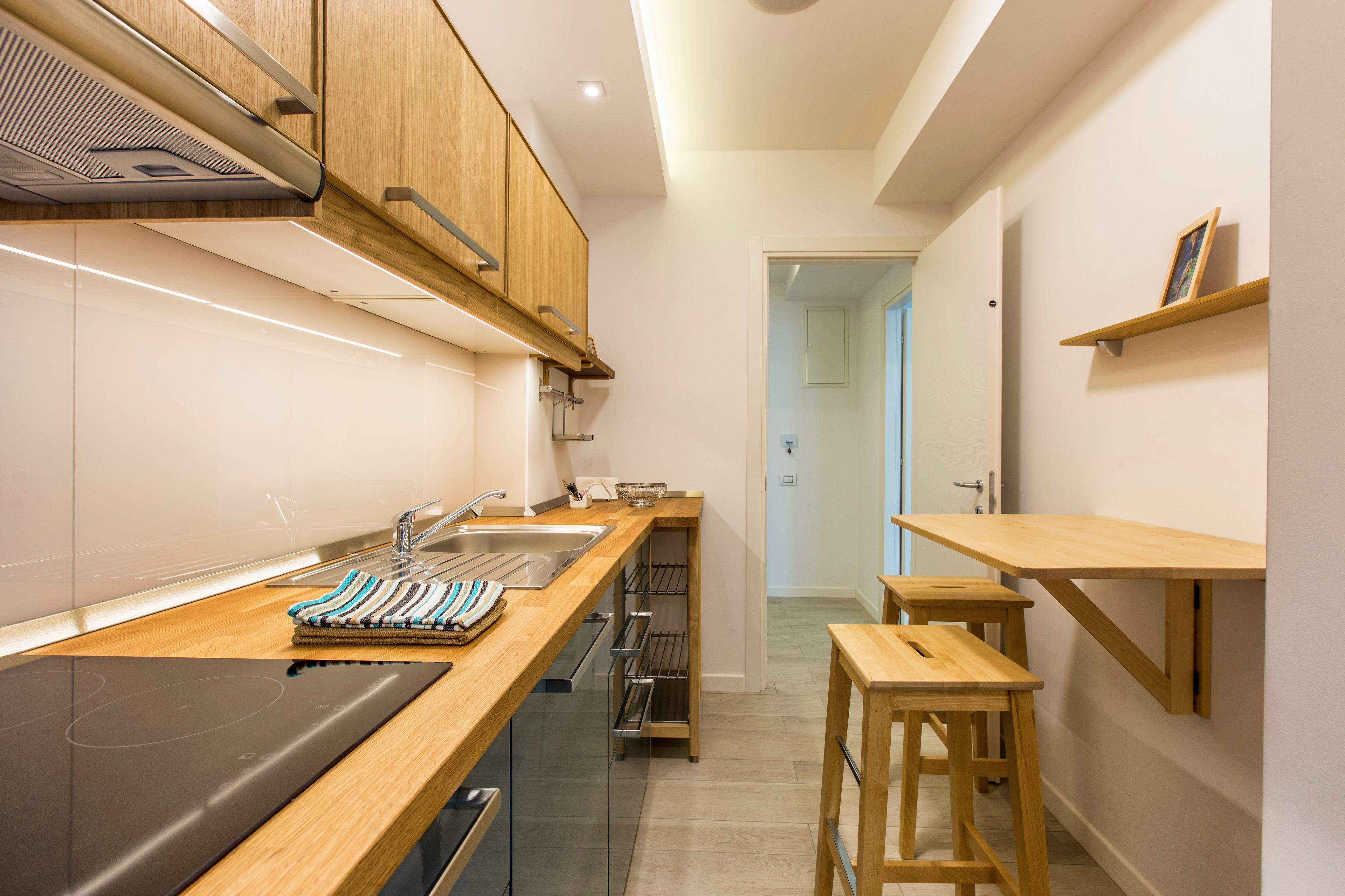 Una cocina moderna con armarios de madera, un lavamanos de acero inoxidable y un horno empotrado. Hay taburetes de madera y una pequeña mesa.