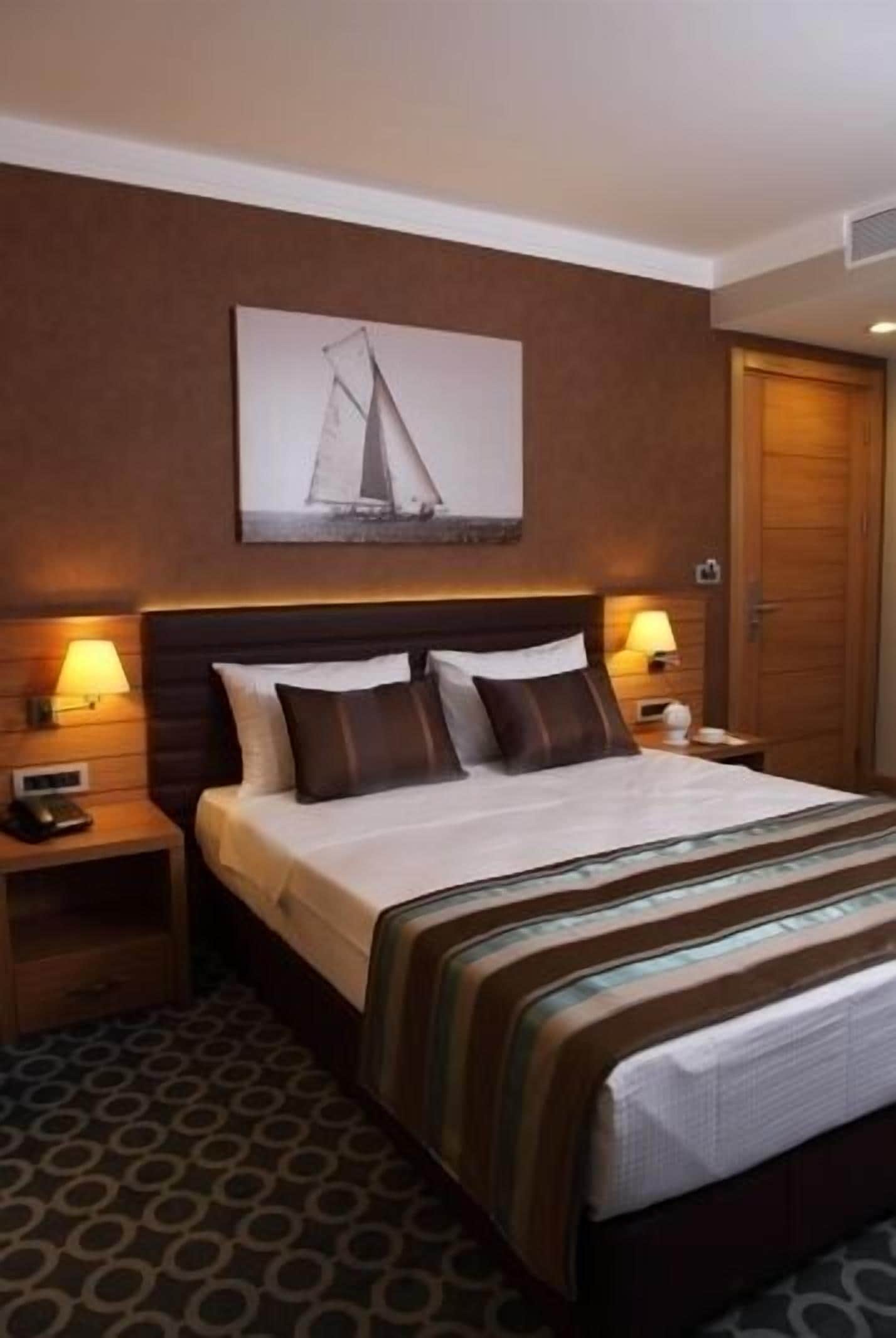 A hotel room with a large bed, two bedside lamps, a wooden headboard, a framed picture on the wall, and a patterned rug.
