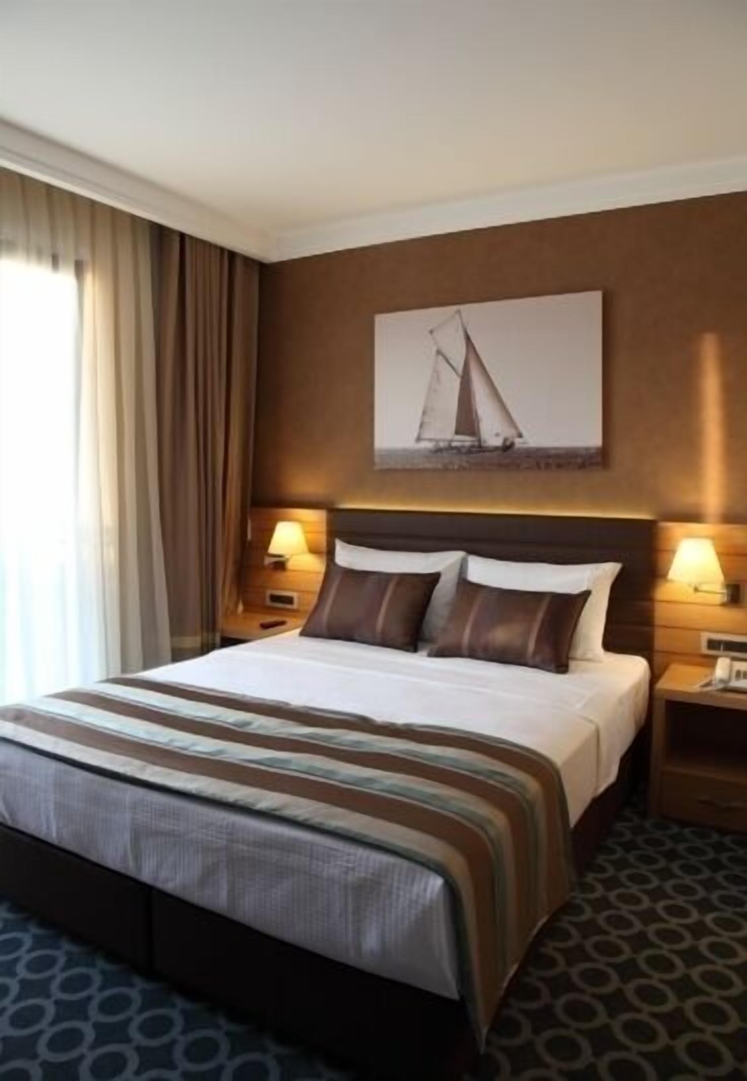 A hotel room with a large bed, two bedside tables with lamps, a headboard with a sailboat painting, and a window with curtains.