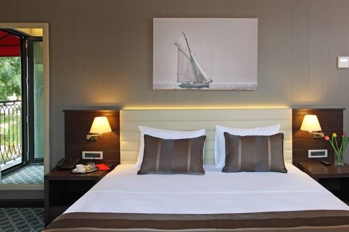 A hotel room with a large bed, two bedside tables with lamps, a painting on the wall, and a view through the door.