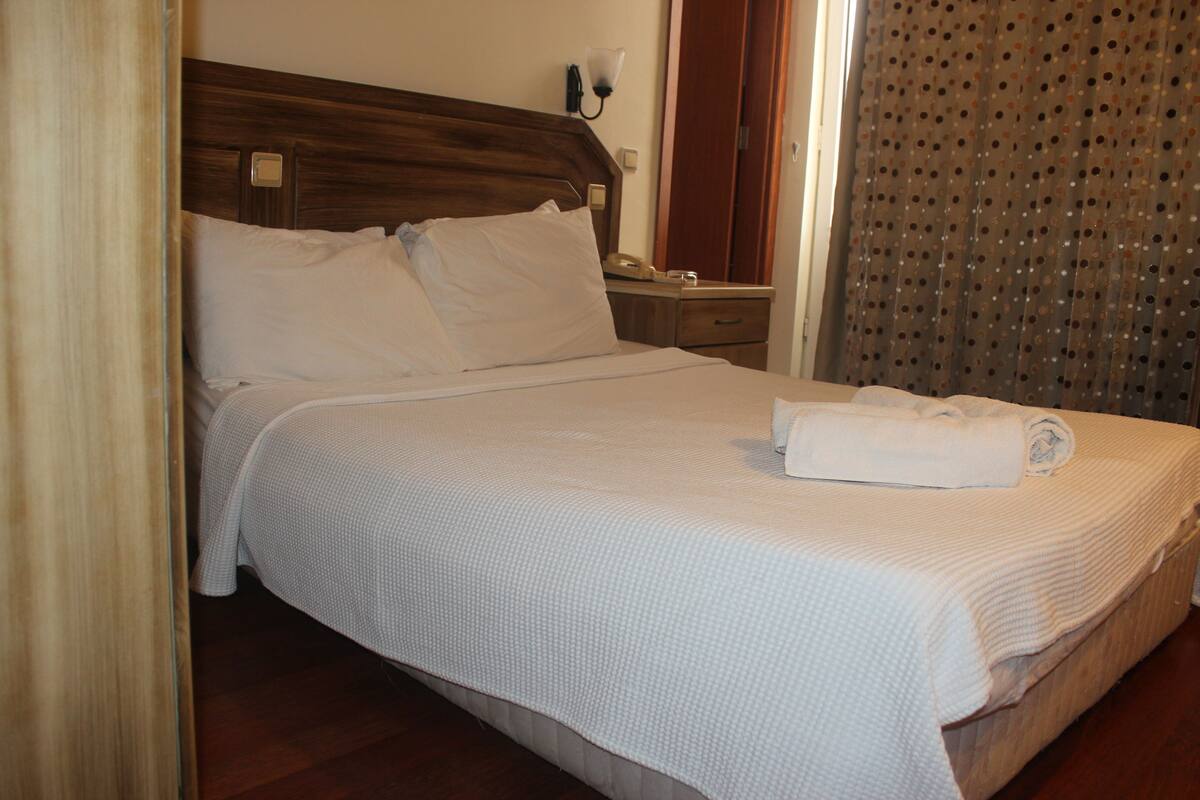 A neatly made bed with a white bedspread and pillows, a wooden headboard, a bedside table with a lamp, and a curtain with a dotted pattern.