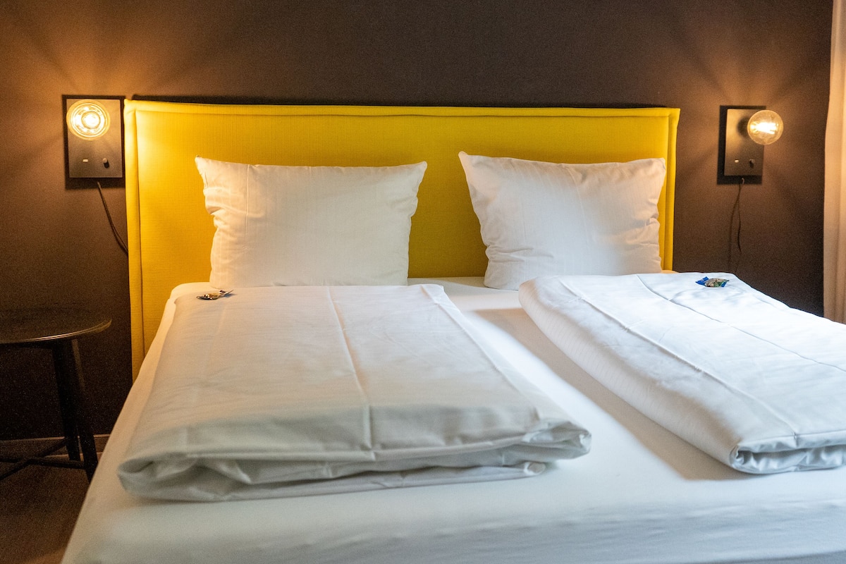 A hotel room with a double bed, two pillows, a bedside table, and two wall-mounted lamps.