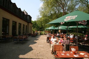 An outdoor café with tables and chairs, green umbrellas, and a building with large windows. - Hotel Haus Hohenstein (Witten)