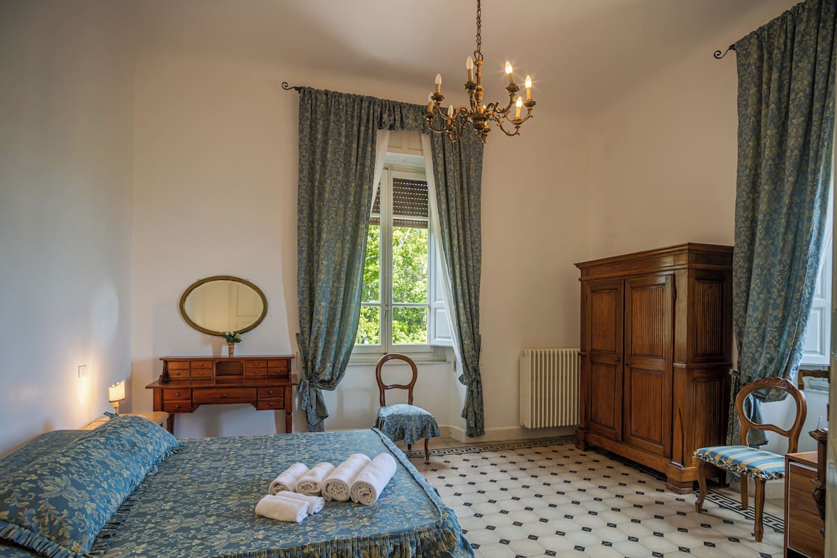 Quarto com cama, guarda-roupa de madeira, cadeira, penteadeira, espelho e janela com cortinas.