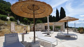 A sunlit outdoor area with lounge chairs and thatched umbrellas, stone walls, and a view of trees.