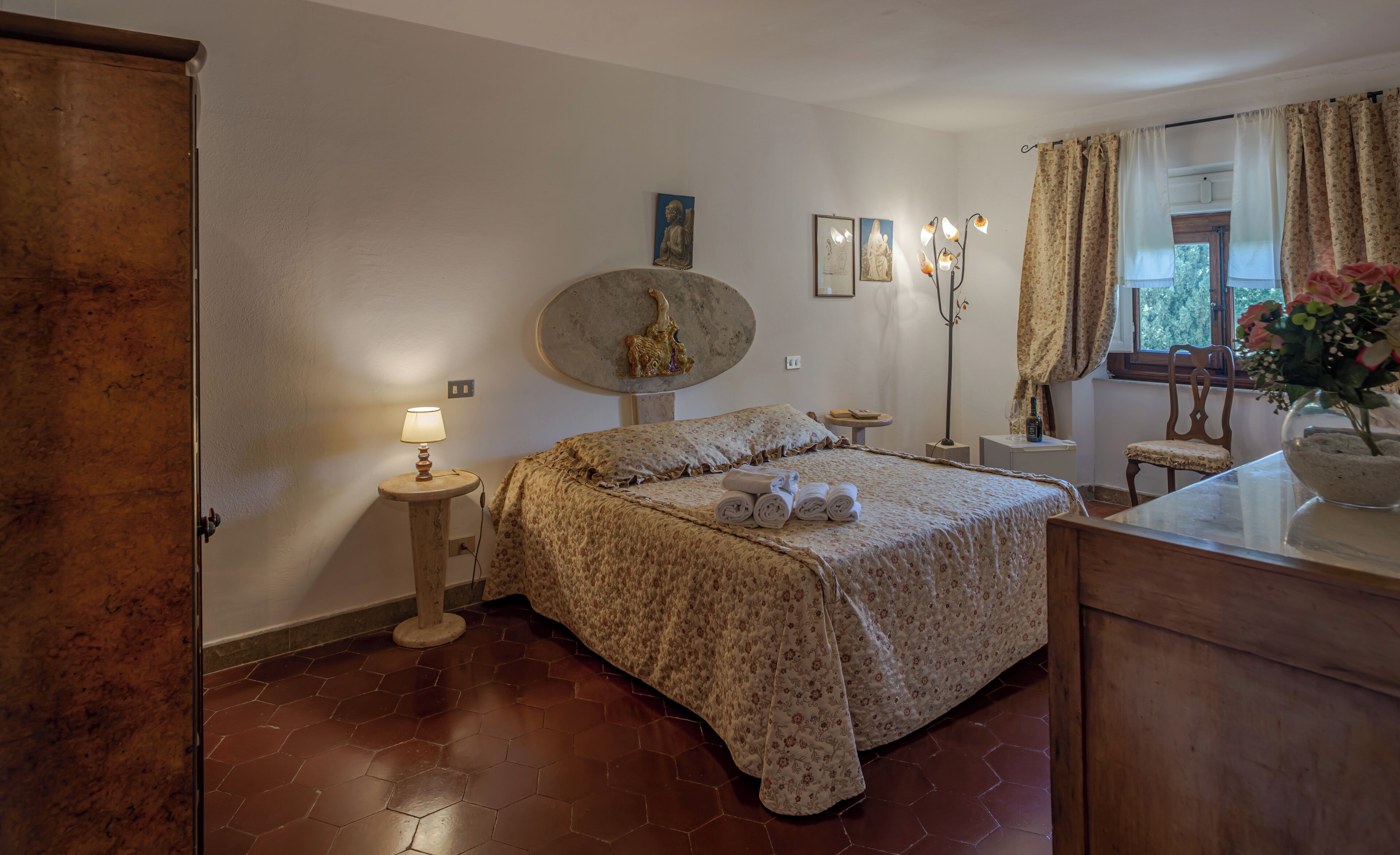 A bedroom with a bed, a nightstand, a lamp, a vase with flowers, and a window with curtains.