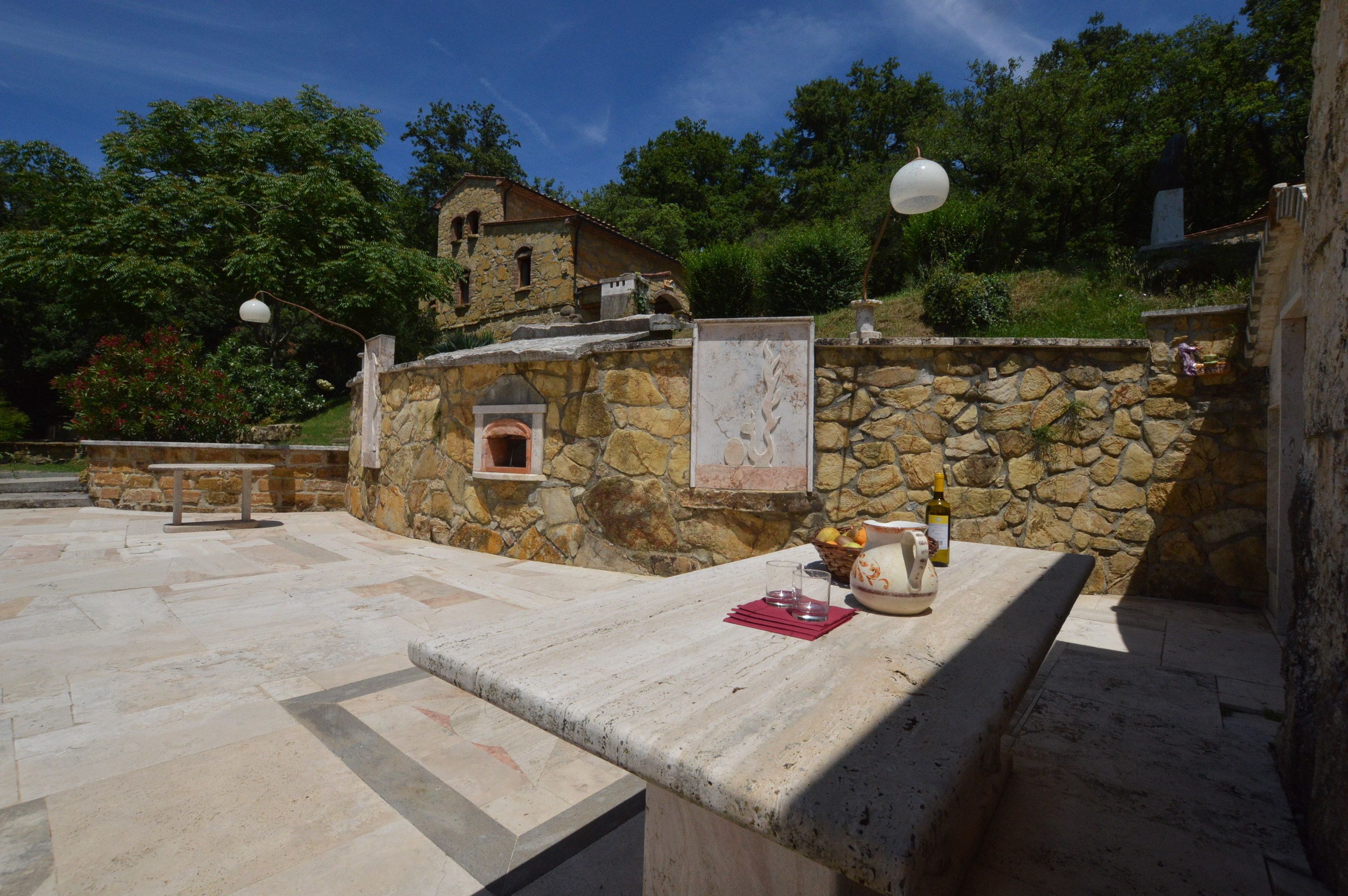 A stone-walled outdoor area with a stone bench, a table with a bottle and glasses, and a view of a building in the background.