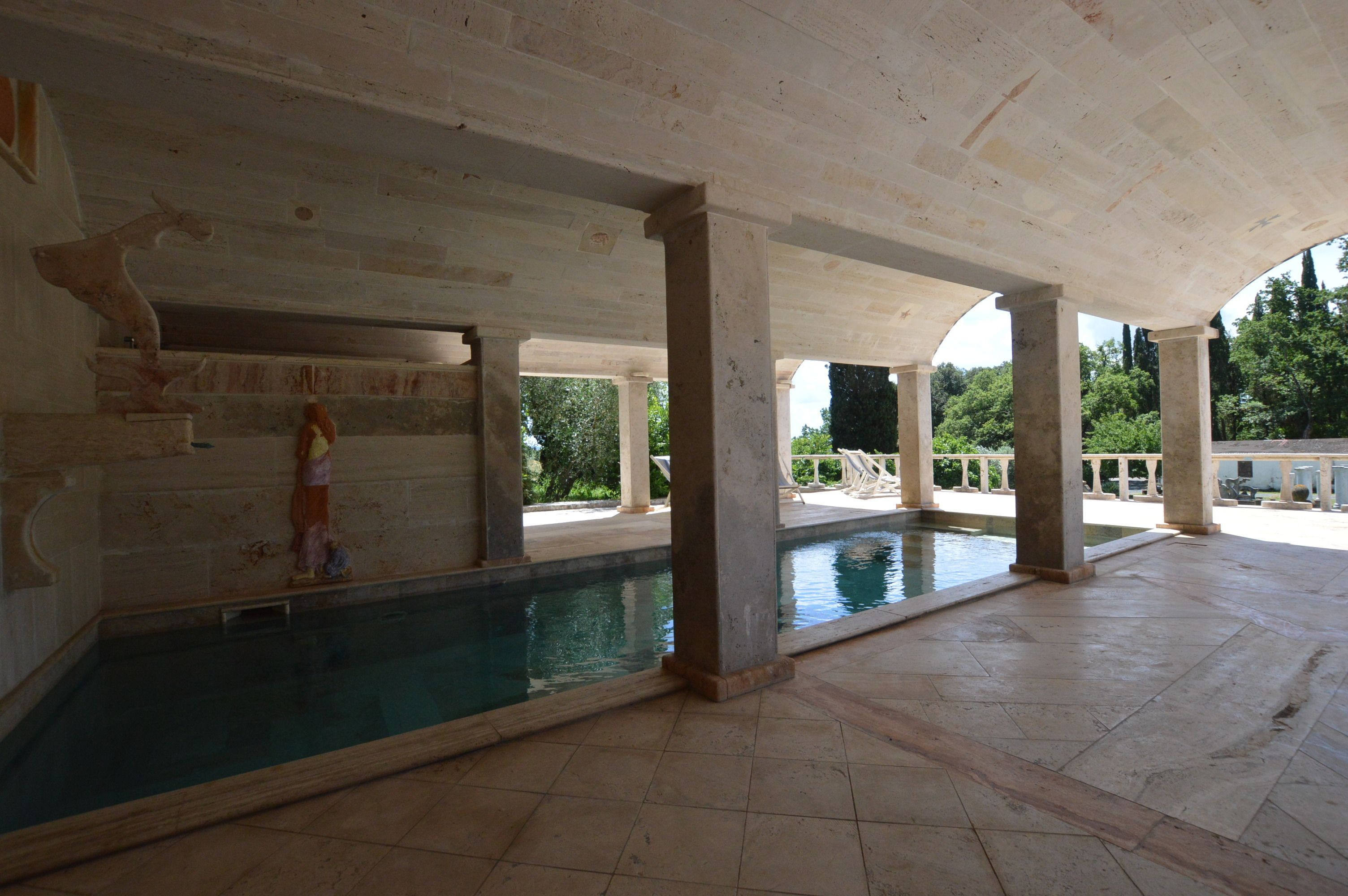 A large indoor swimming pool with a high ceiling and columns, surrounded by a spacious area with a view of greenery outside.