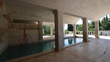 A large indoor swimming pool with a high ceiling and columns, surrounded by a spacious area with a view of greenery outside.
