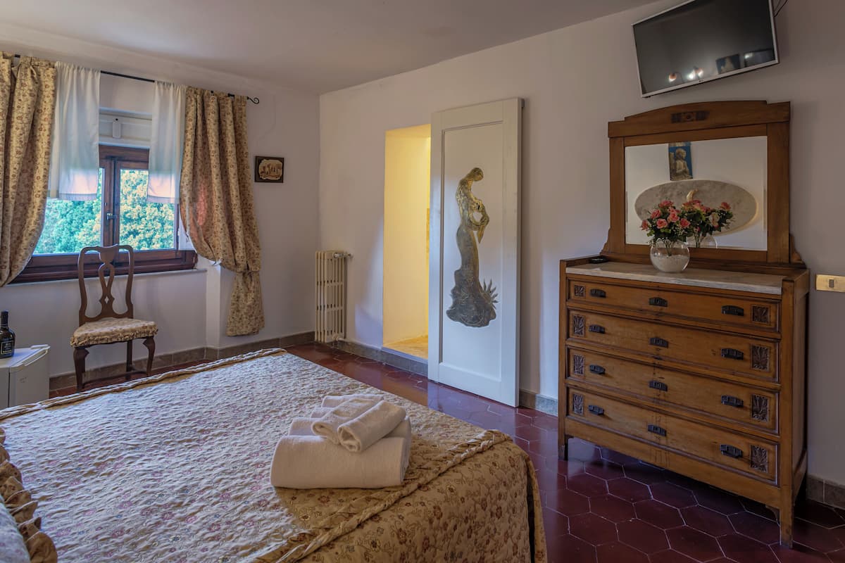 Quarto com cama, cómoda de madeira, cadeira, televisão e janela com cortinas.
