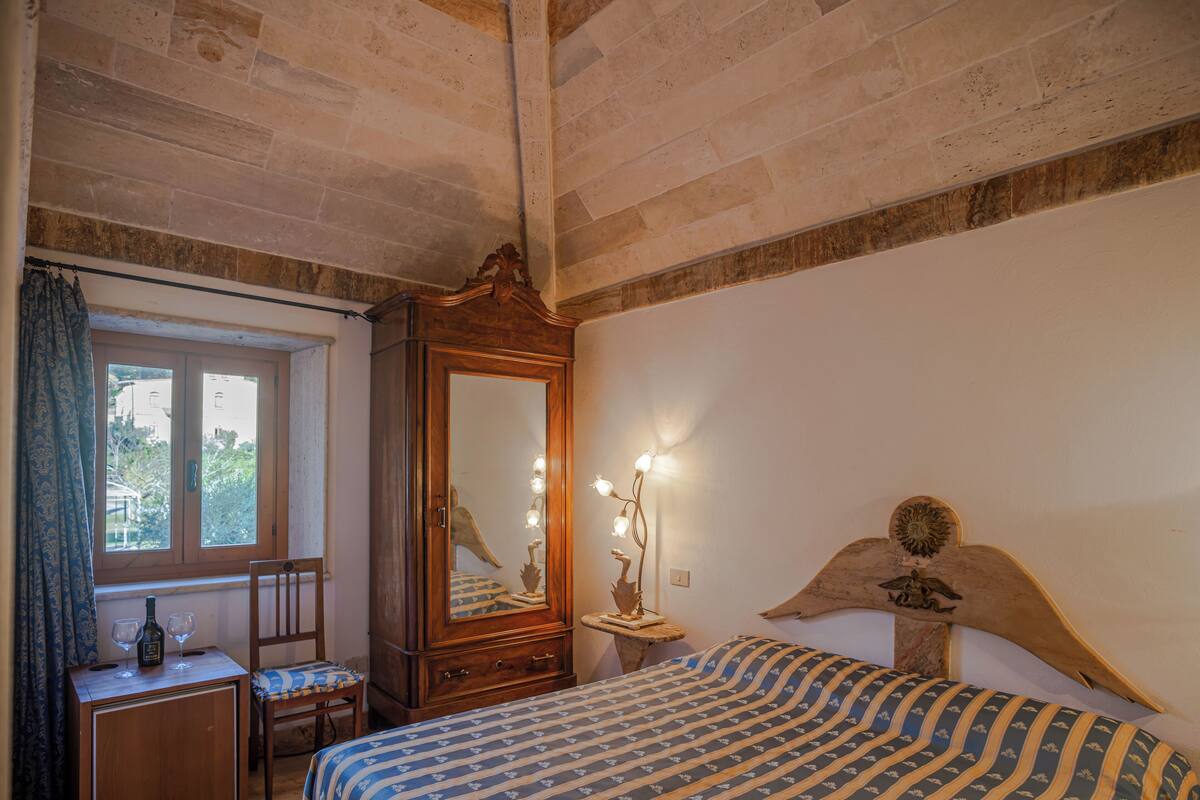 Quarto com cama de madeira, mesa de cabeceira com abajur, cômoda e janela com cortinas.