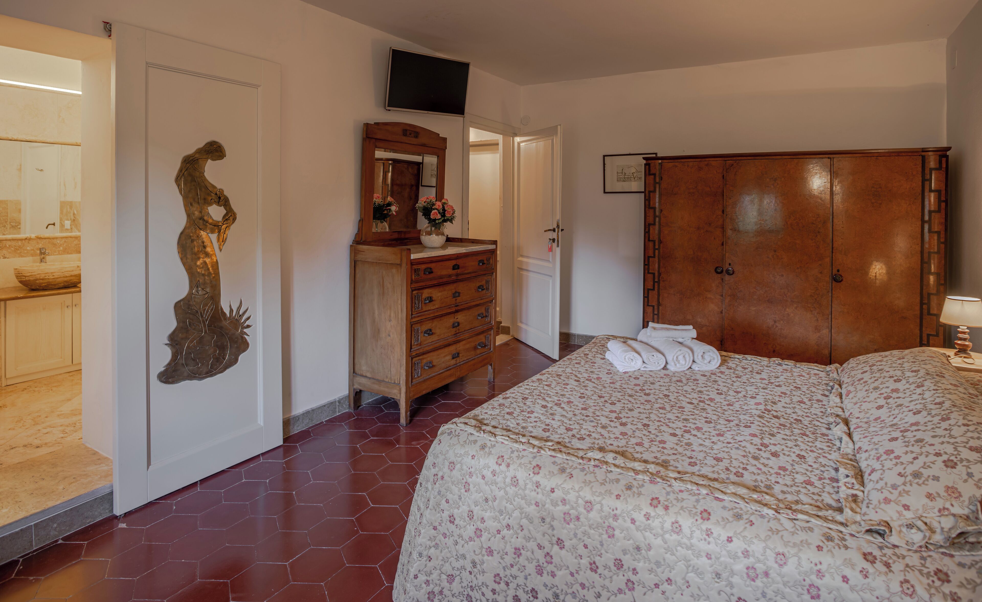 A bedroom with a wooden wardrobe, a bed with floral bedding, a dresser, and a wall-mounted TV.