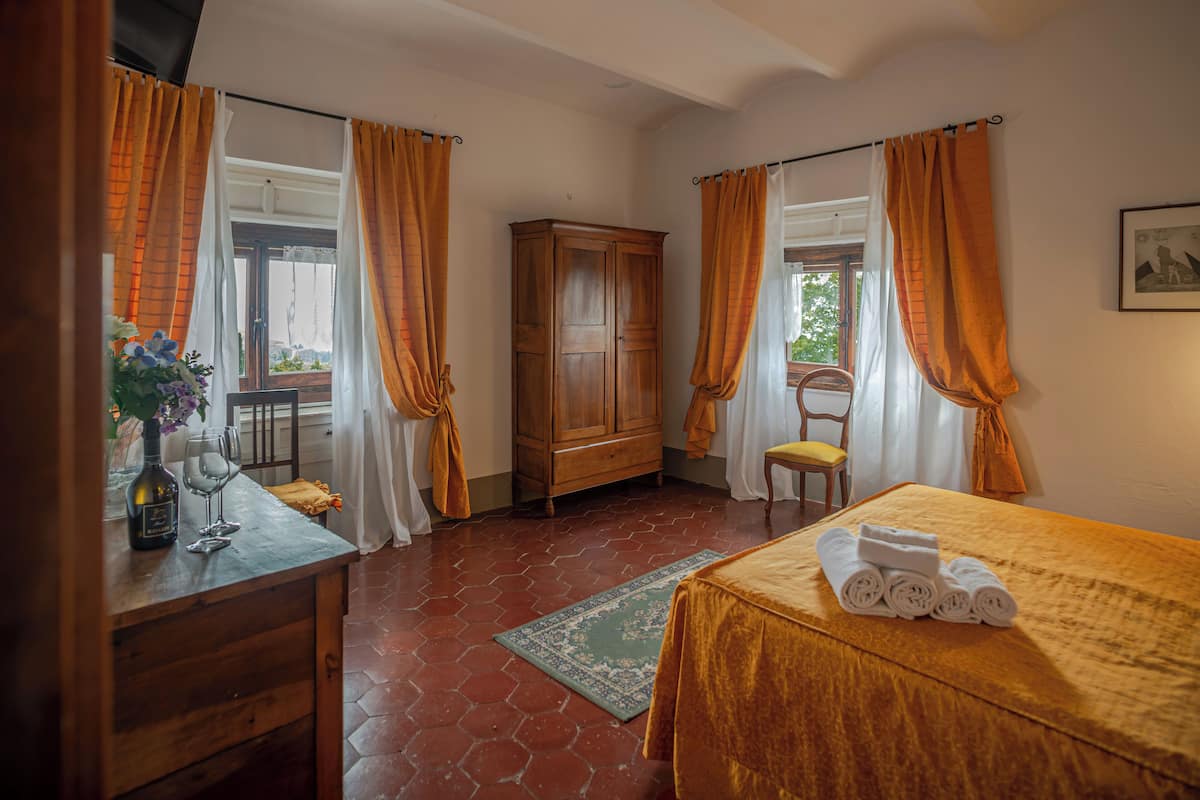 Quarto de hotel com cama, um armário de madeira, uma cadeira, mesa com um vaso e copos de vinho, e cortinas laranja.