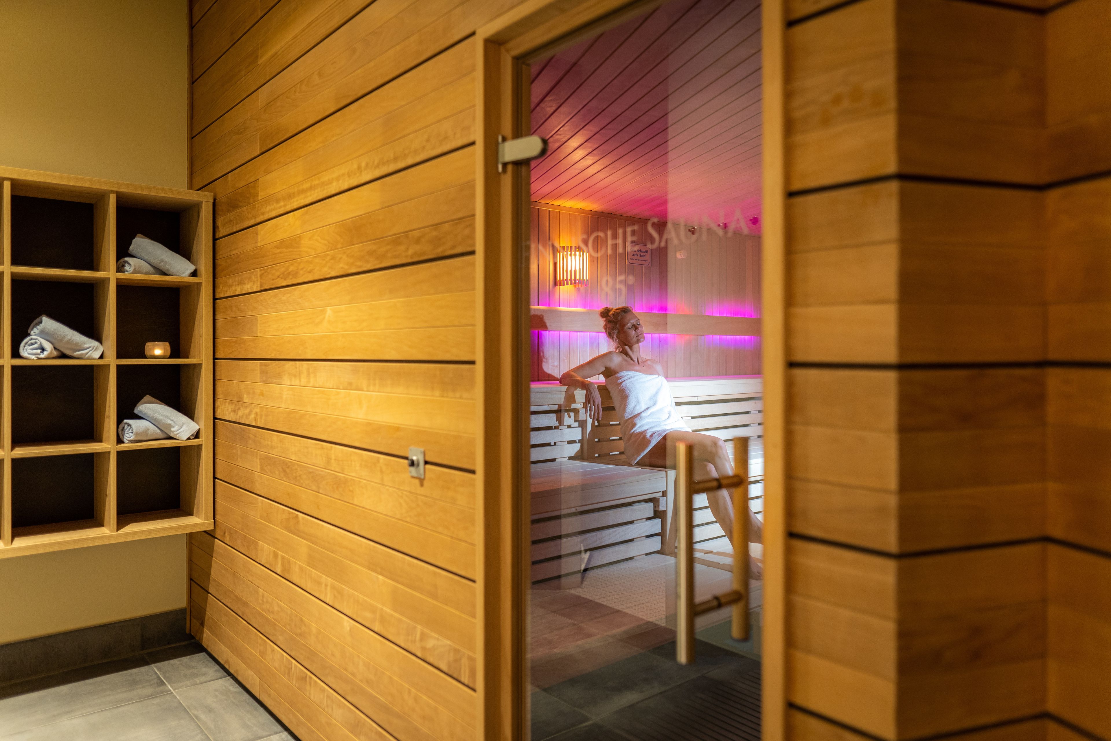 A wooden sauna room with a glass door, shelves with towels, and a bench.