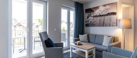 Un salón moderno con un sofá gris, dos sillones, una mesa de centro y un ventanal con vistas a casas.