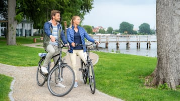 Dos personas en bicicleta sobre un sendero de grava, con un lago y árboles al fondo.