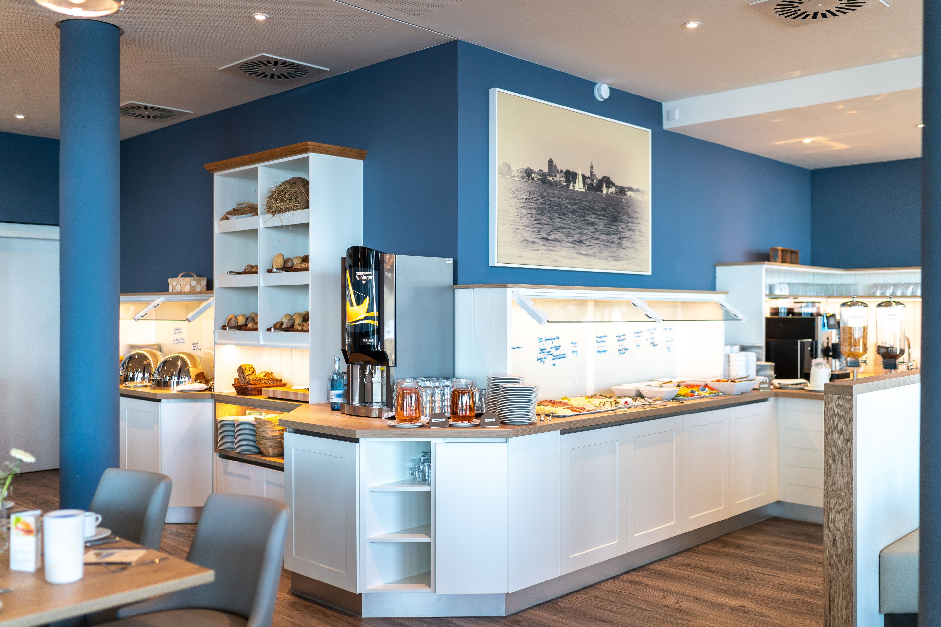 A modern cafe with a blue and white color scheme, featuring a counter with food and drinks, a coffee machine, and a cityscape artwork on the wall.
