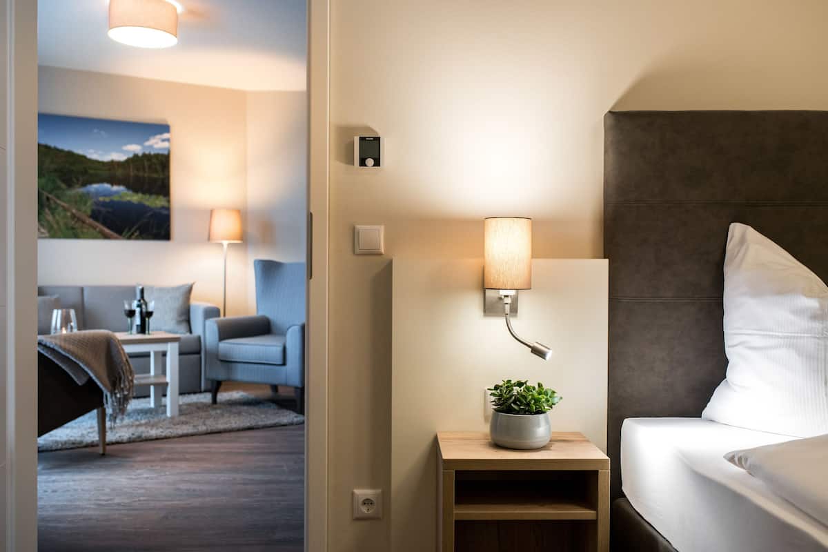 Quarto de hotel moderno com cama, mesa de cabeceira com abajur, uma pequena planta e um quadro na parede.