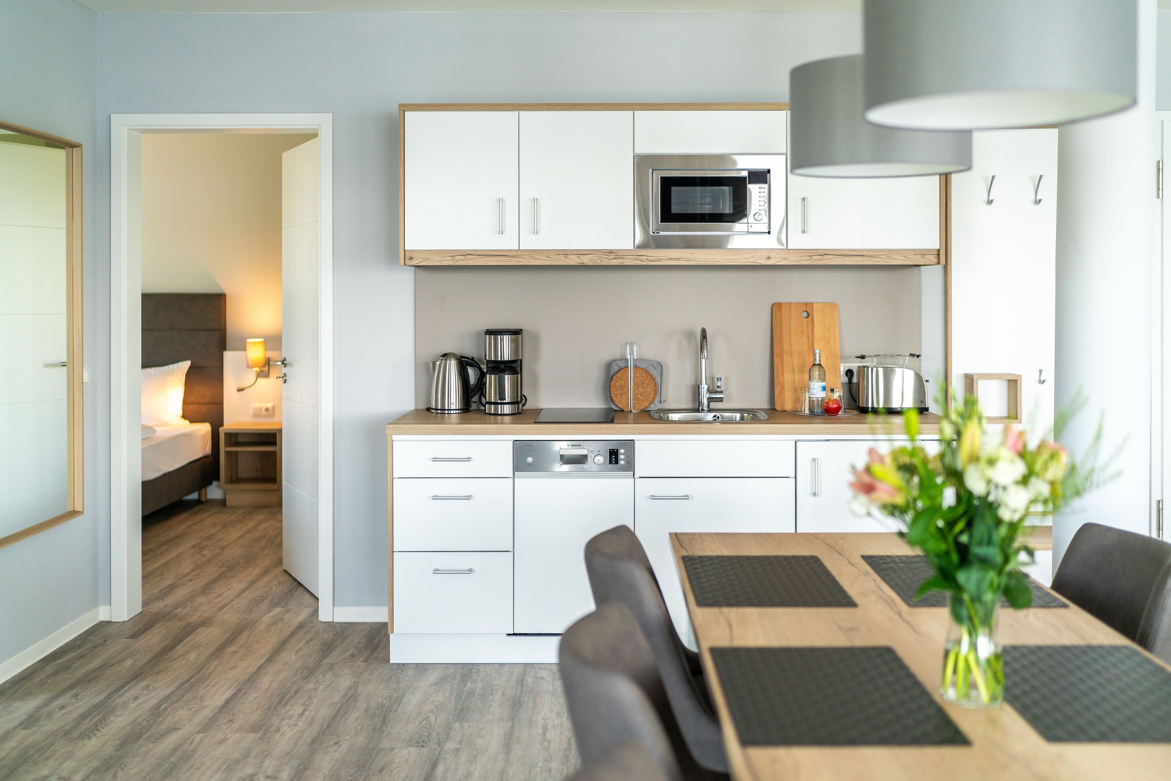A modern kitchen with white cabinets, a microwave, and a coffee maker. There is a dining table with chairs and a vase of flowers.