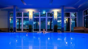 Une grande piscine intérieure aux parois en verre transparent, offrant une vue sur les arbres à l’extérieur, et une personne se relaxant dans l’eau.
