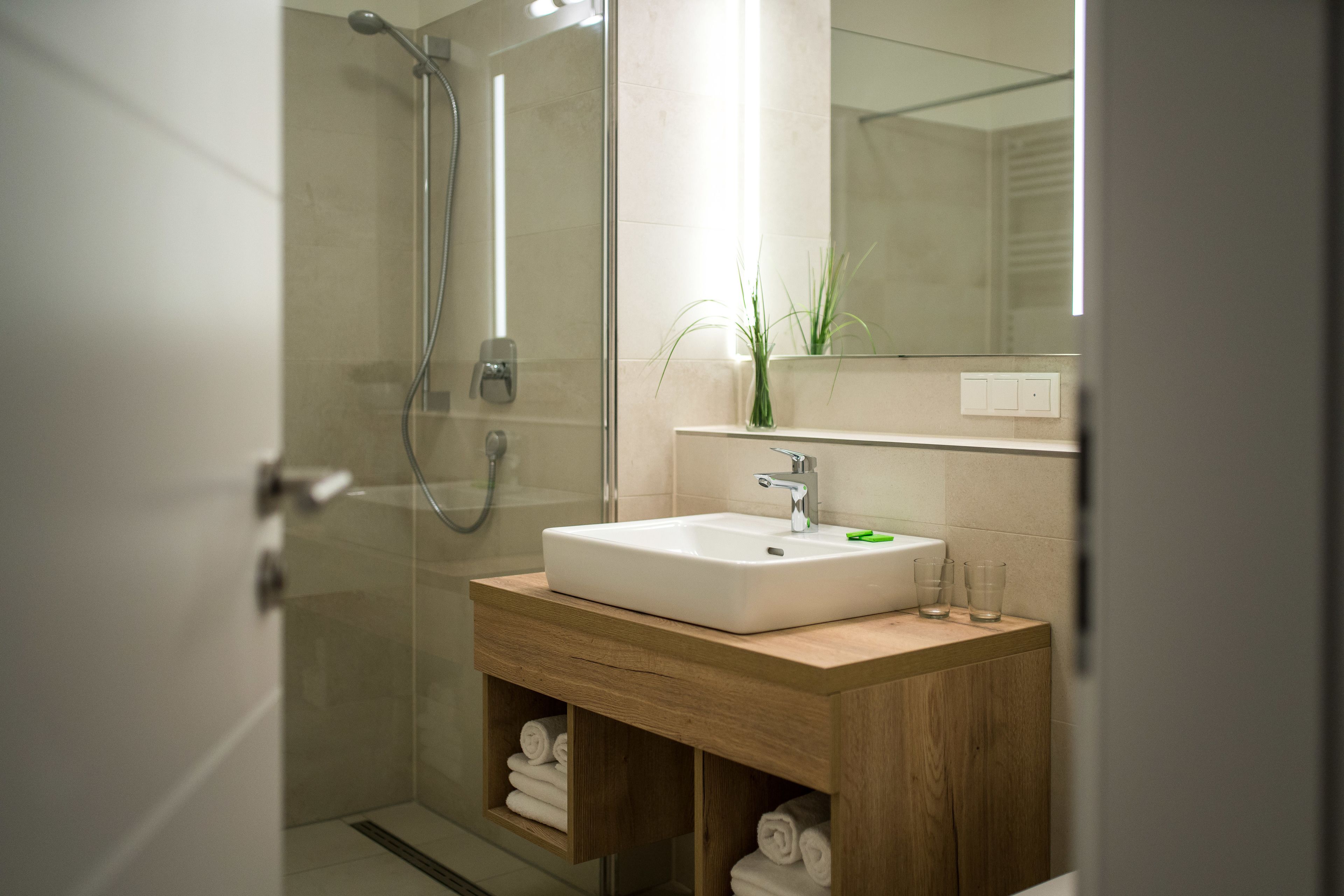 Un baño moderno con ducha de vidrio, tocador de madera con lavamanos blanco y un espejo sobre el lavamanos.