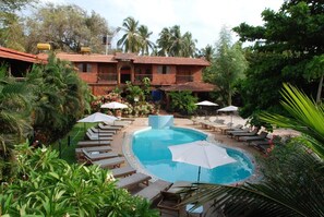 A pool area with lounge chairs and umbrellas, surrounded by lush greenery and a building with a tiled roof. - Sea Breeze Beach Resort Candolim (Candolim)