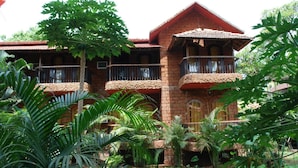 A two-story building with a red-tiled roof, balconies with metal railings, and a garden with various tropical plants.