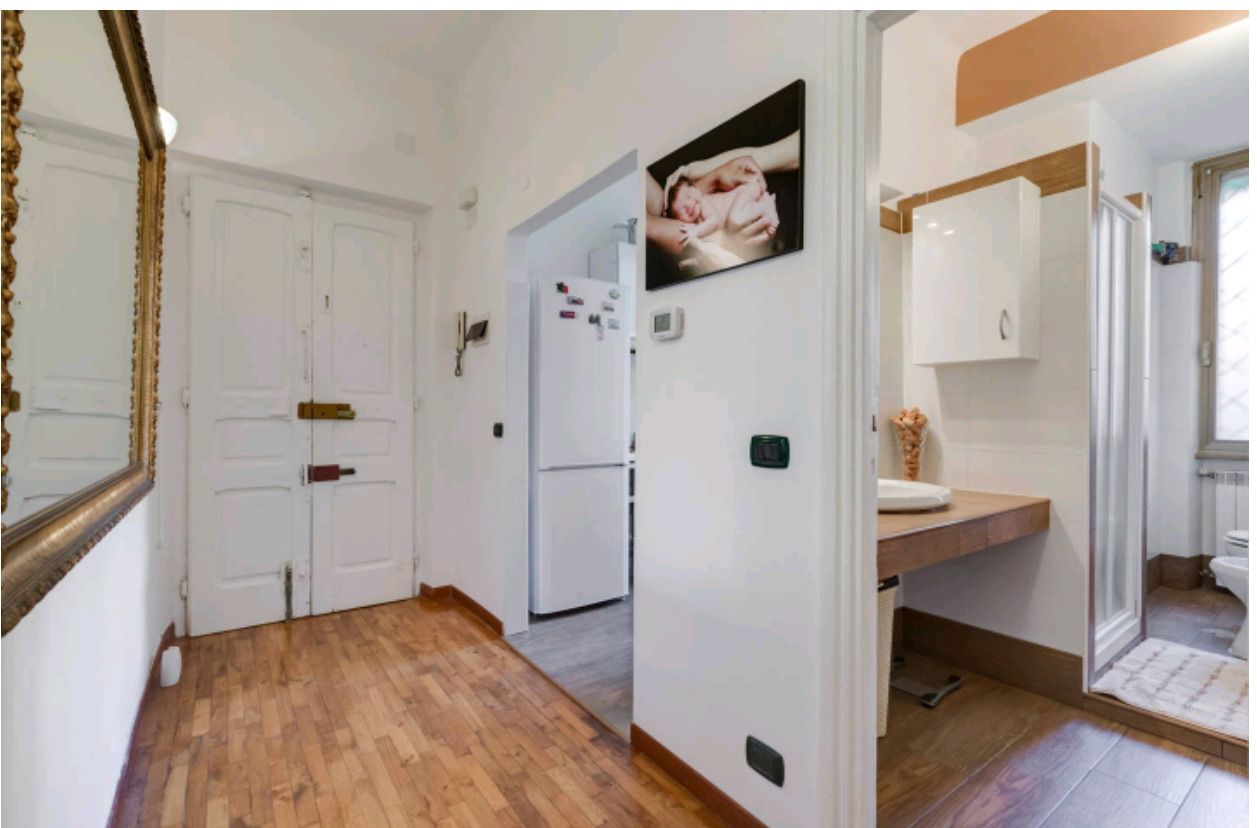 A hallway with wooden flooring, white walls, and a door leading to a kitchen area with a refrigerator and a microwave.
