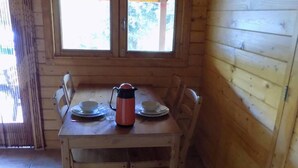 Puisesta ruokapöydästä ja tuoleista koostuva ruokailuryhmä, vedenkeitin ja lautaset. Huoneessa on ikkunat, joissa on verhot, ja seinällä on taulu.