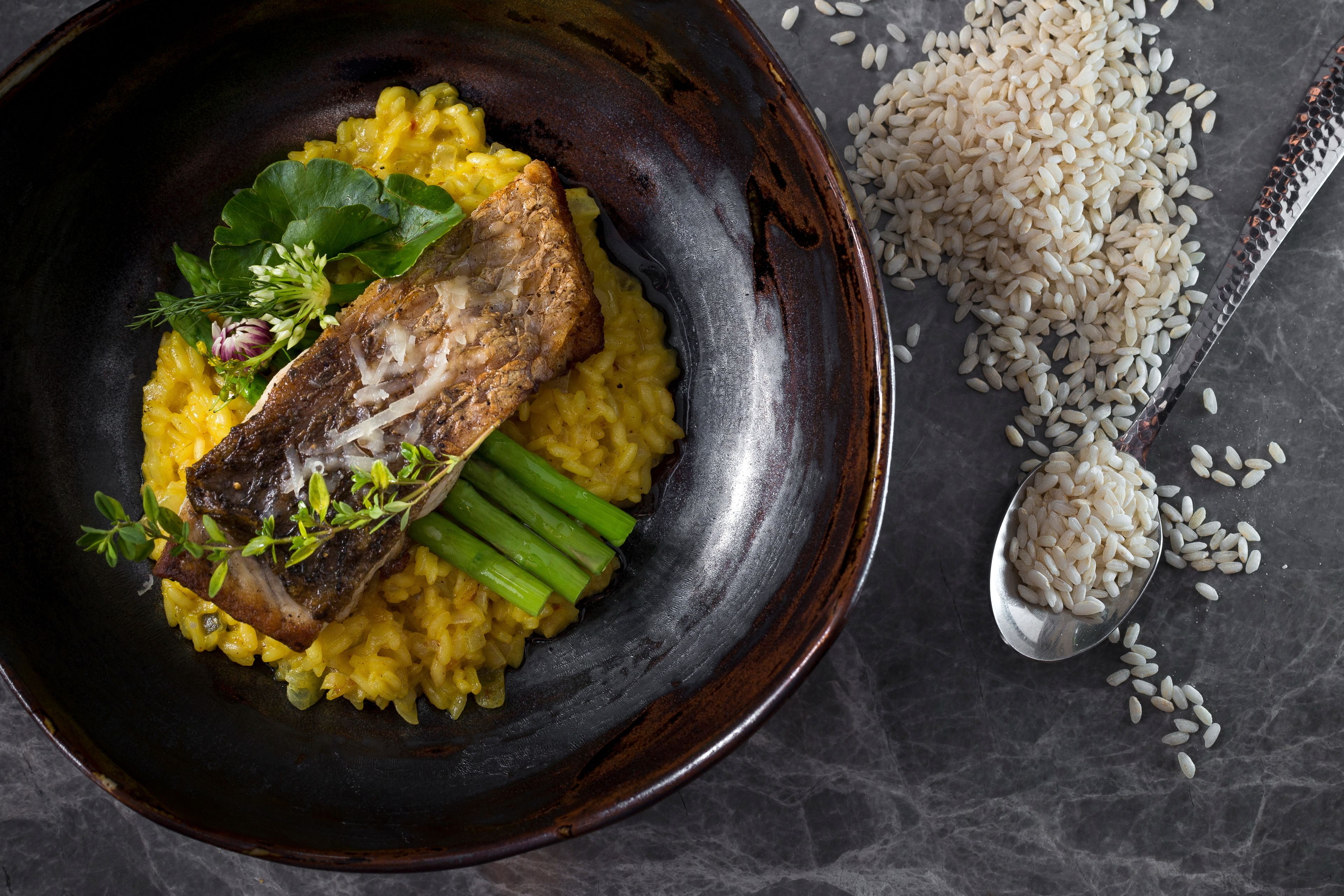 Un plato de comida con arroz, judías verdes y un trozo de carne, acompañado de una cucharada de arroz y granos de arroz esparcidos sobre una superficie oscura.