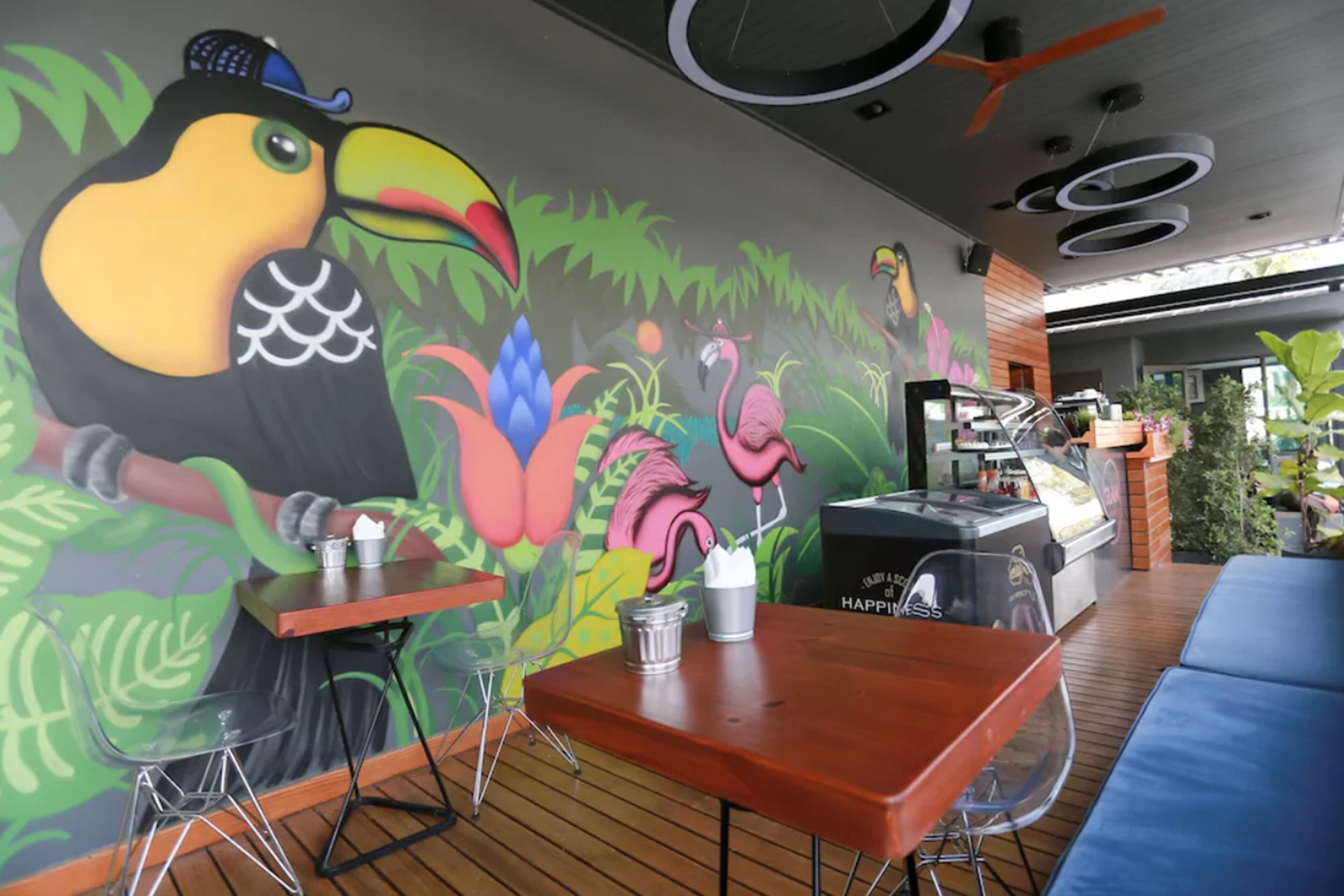 Un restaurante con un mural de un tucán y flamencos, mesas de madera y una zona de parilla.