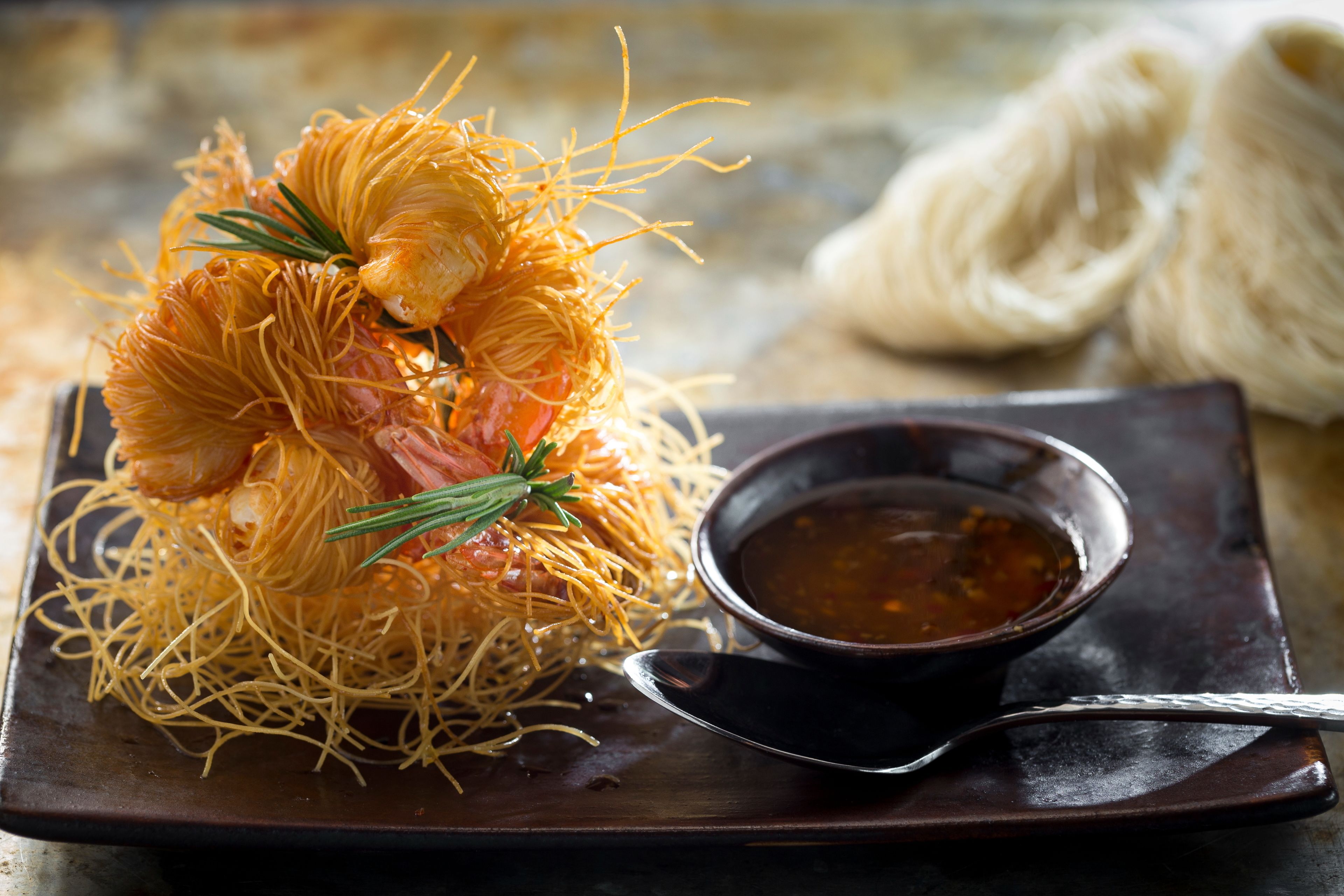 Un plat de crevettes enroulées dans des nouilles, garni d’une branche de romarin, servi dans une assiette sombre avec un petit bol de sauce et une cuillère.