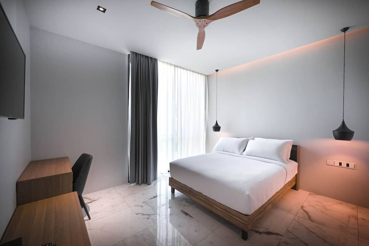 A modern hotel room with a large bed, a desk, a chair, a ceiling fan, and a large window with curtains.