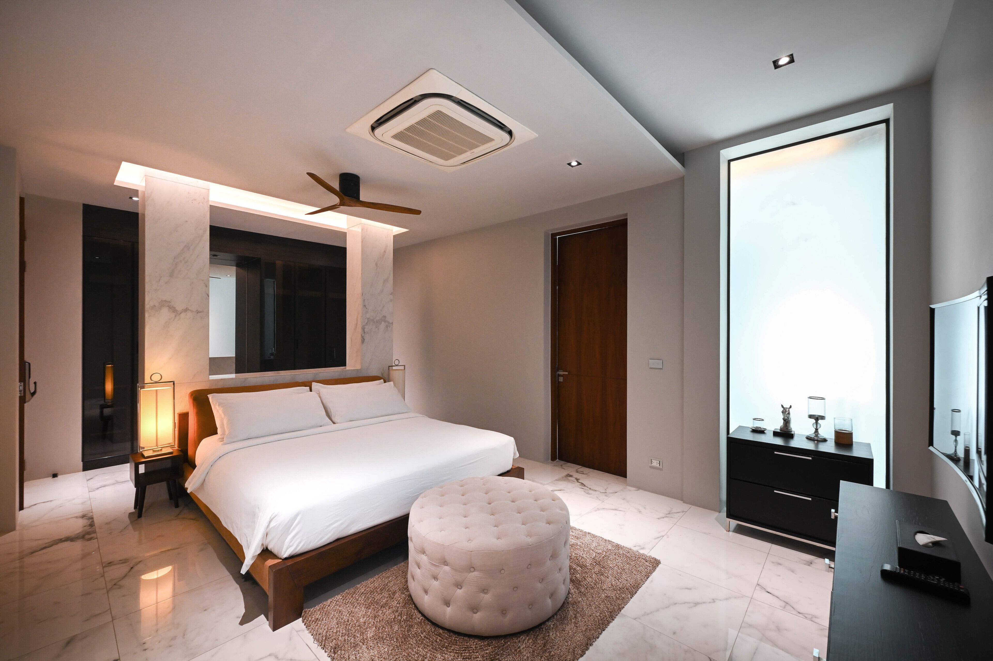 A modern bedroom with a large bed, a round ottoman, a TV, and a wardrobe.