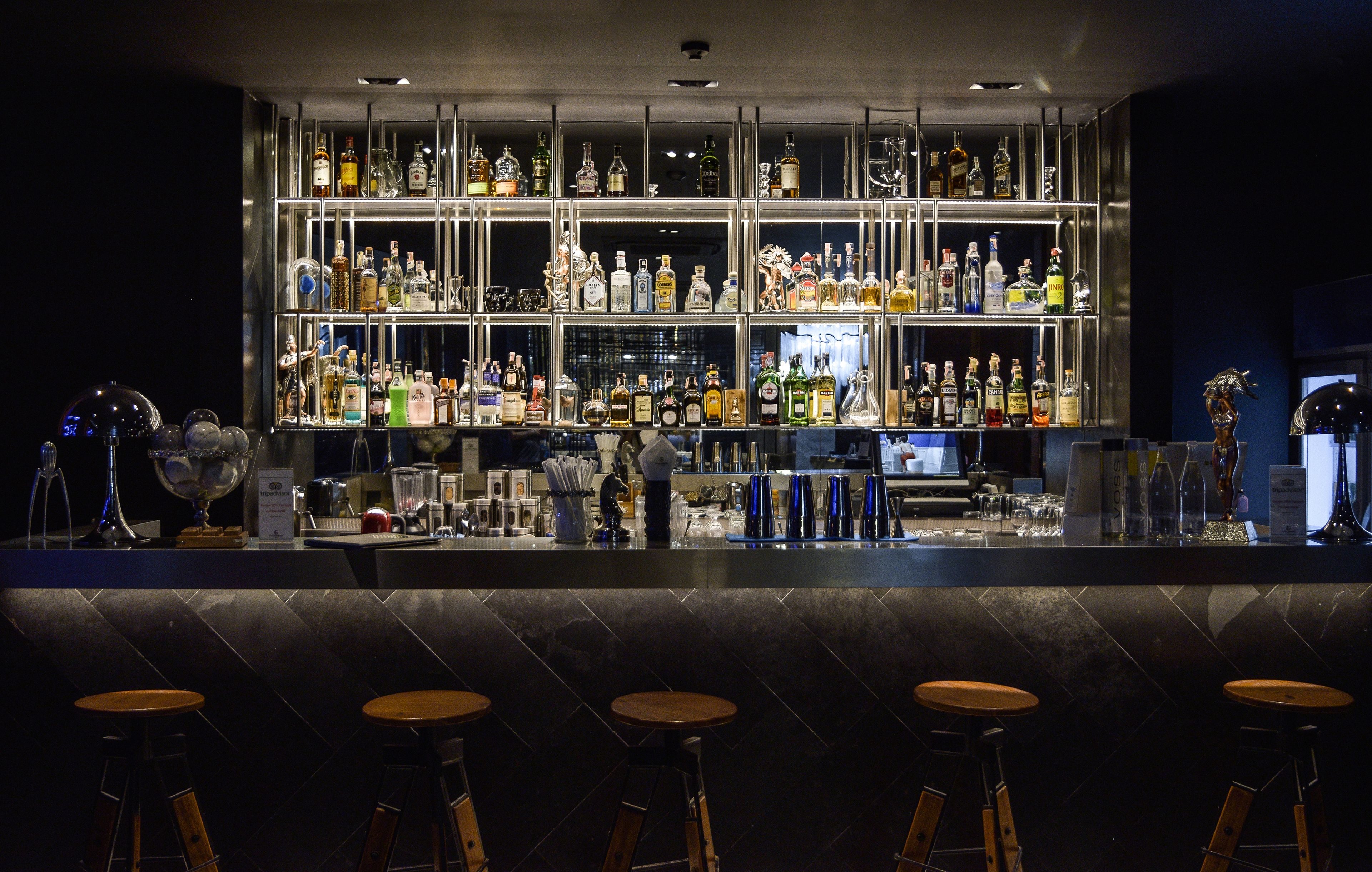 A bar area with multiple shelves of liquor bottles, a bartender, and four wooden bar stools.