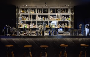 A bar area with multiple shelves of liquor bottles, a bartender, and four wooden bar stools. - Glam Habitat Hotel (Kamala)
